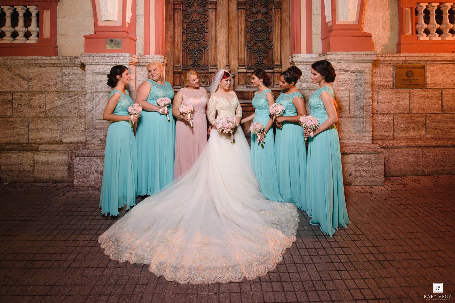 Bride in white gown poses with six bridesmaids in teal dresses outside a historic church building.