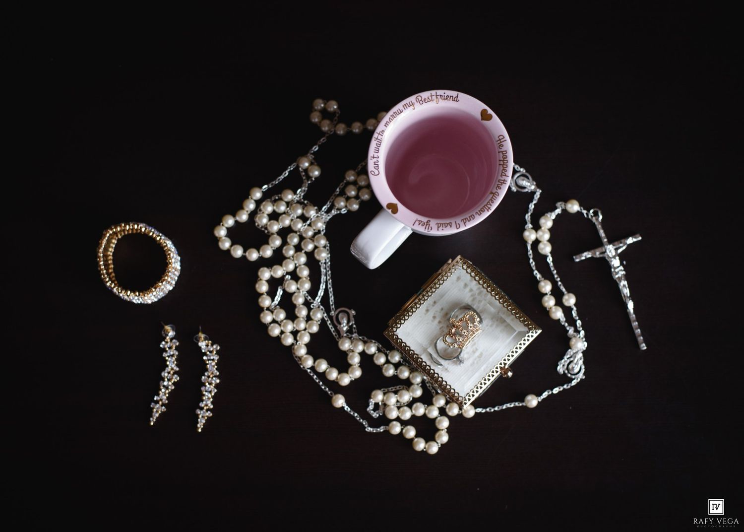 Flat lay of pearl rosary beads, crucifix, gold rings, earrings, and a pink drink in a white mug on black background.