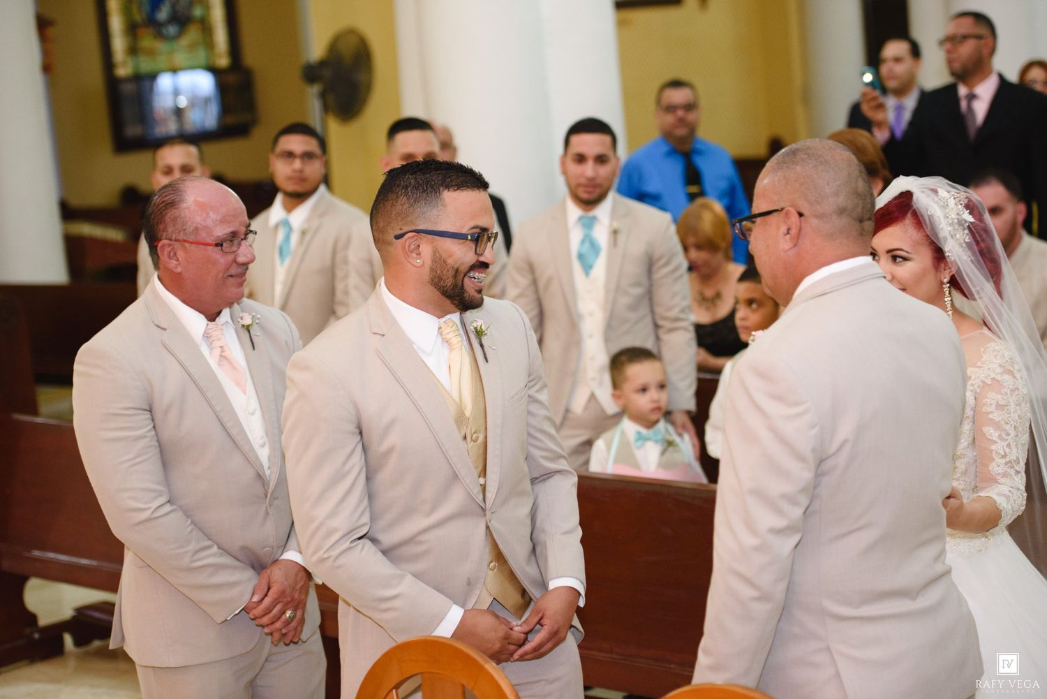 Groom in beige suit greeting guests at church wedding ceremony with bridal party in background.