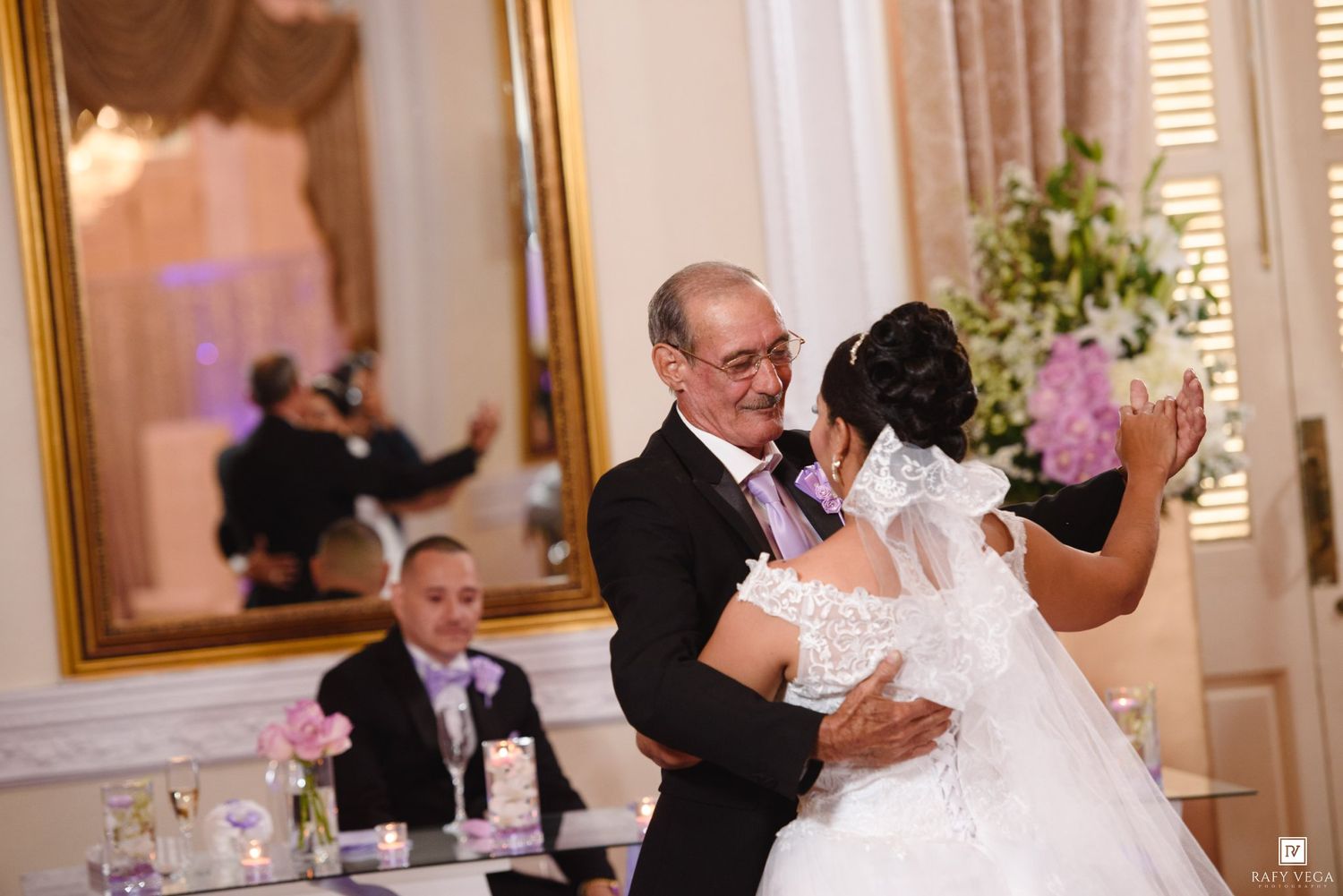 Bride and older man share a father-daughter dance at elegant wedding reception with mirror and floral decor.