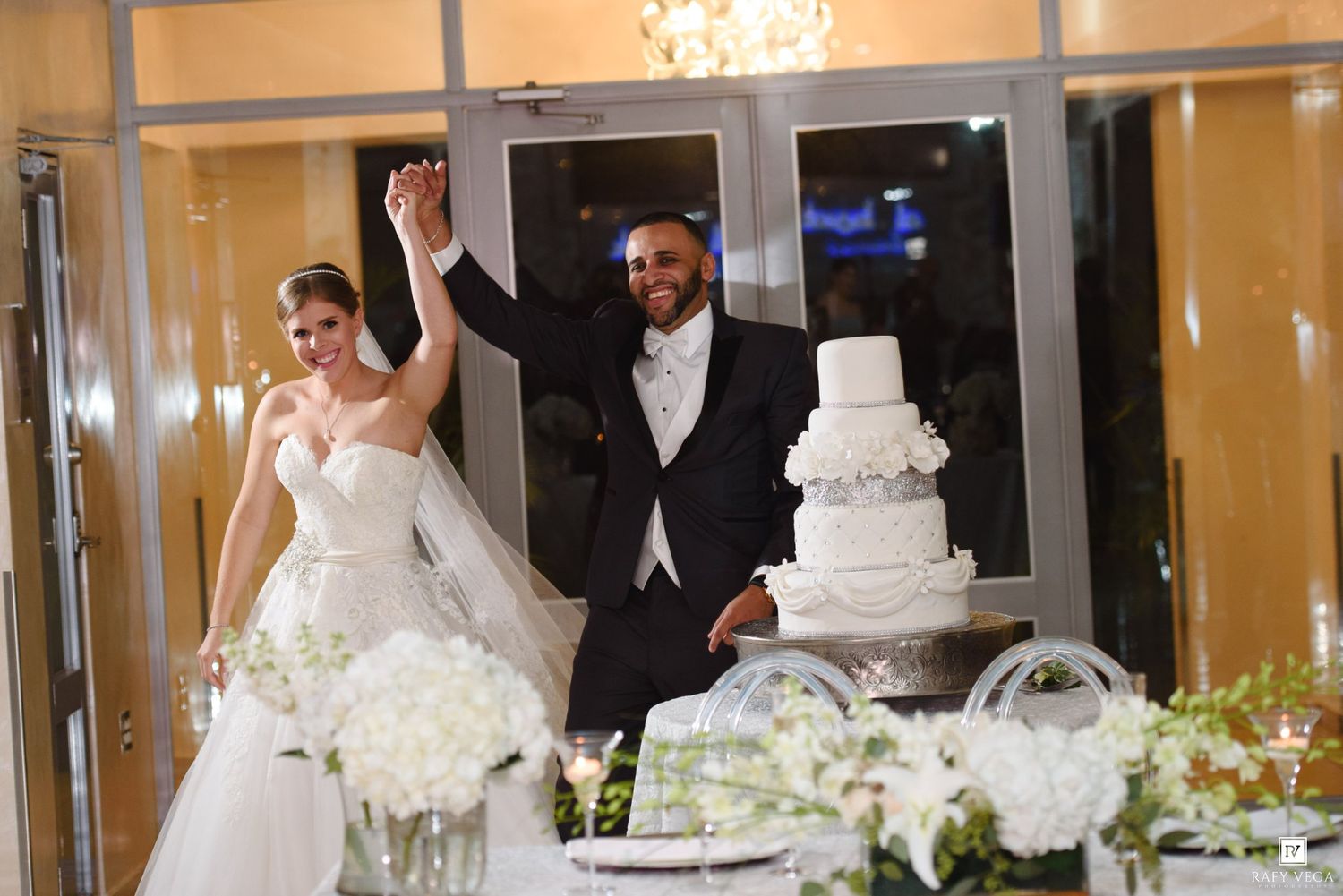 Bride and groom entering wedding reception hall, raising hands in celebration near elegant white tiered cake.