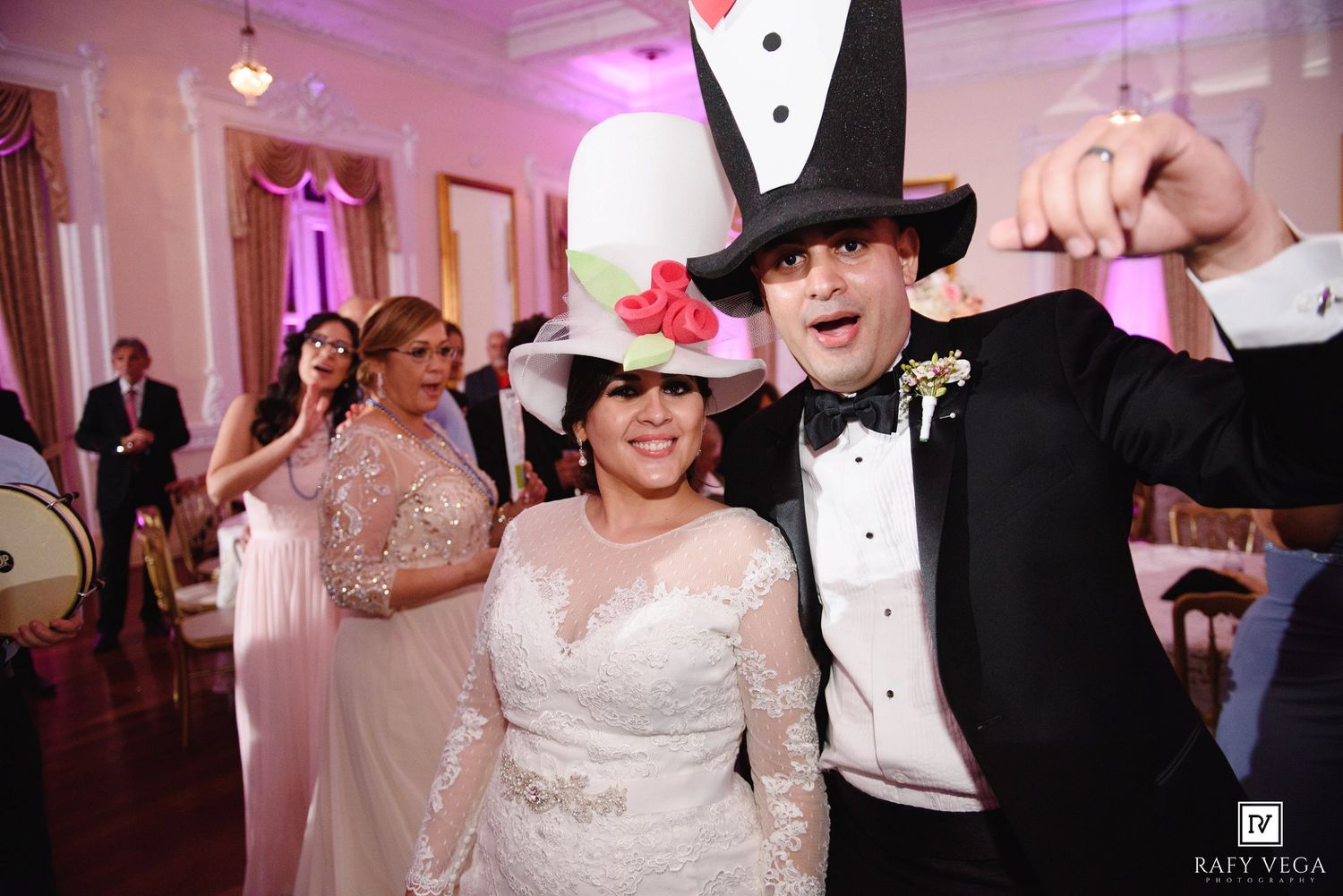 Bride and groom celebrating at wedding reception wearing oversized novelty hats, smiling and dancing in elegant ballroom.