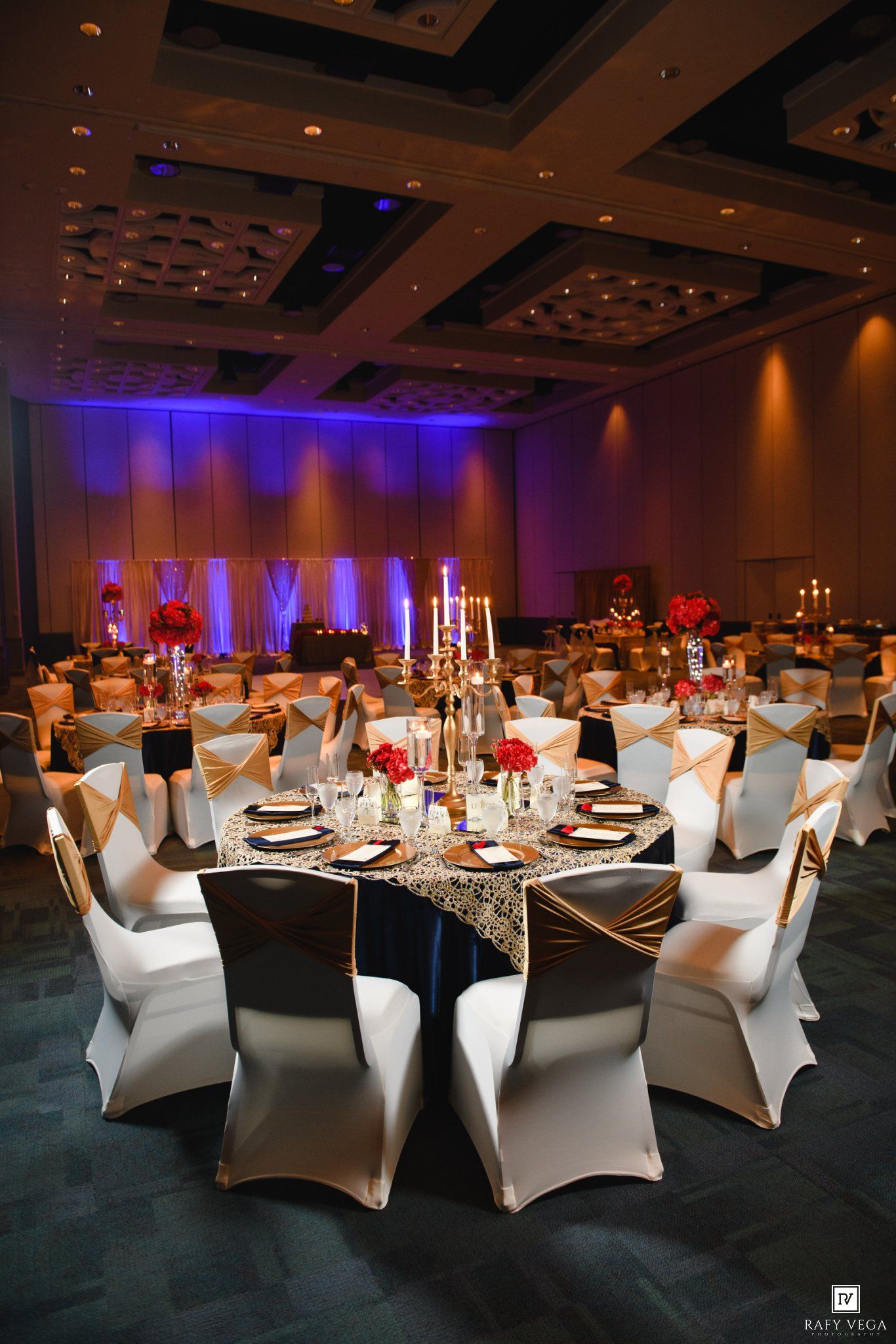 Elegant ballroom reception with white chair covers, gold sashes, black tablecloths, red floral centerpieces, and purple uplighting.