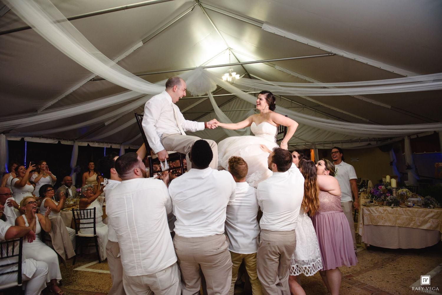 Bride and groom lifted by guests during joyful hora dance at elegant tented wedding reception.