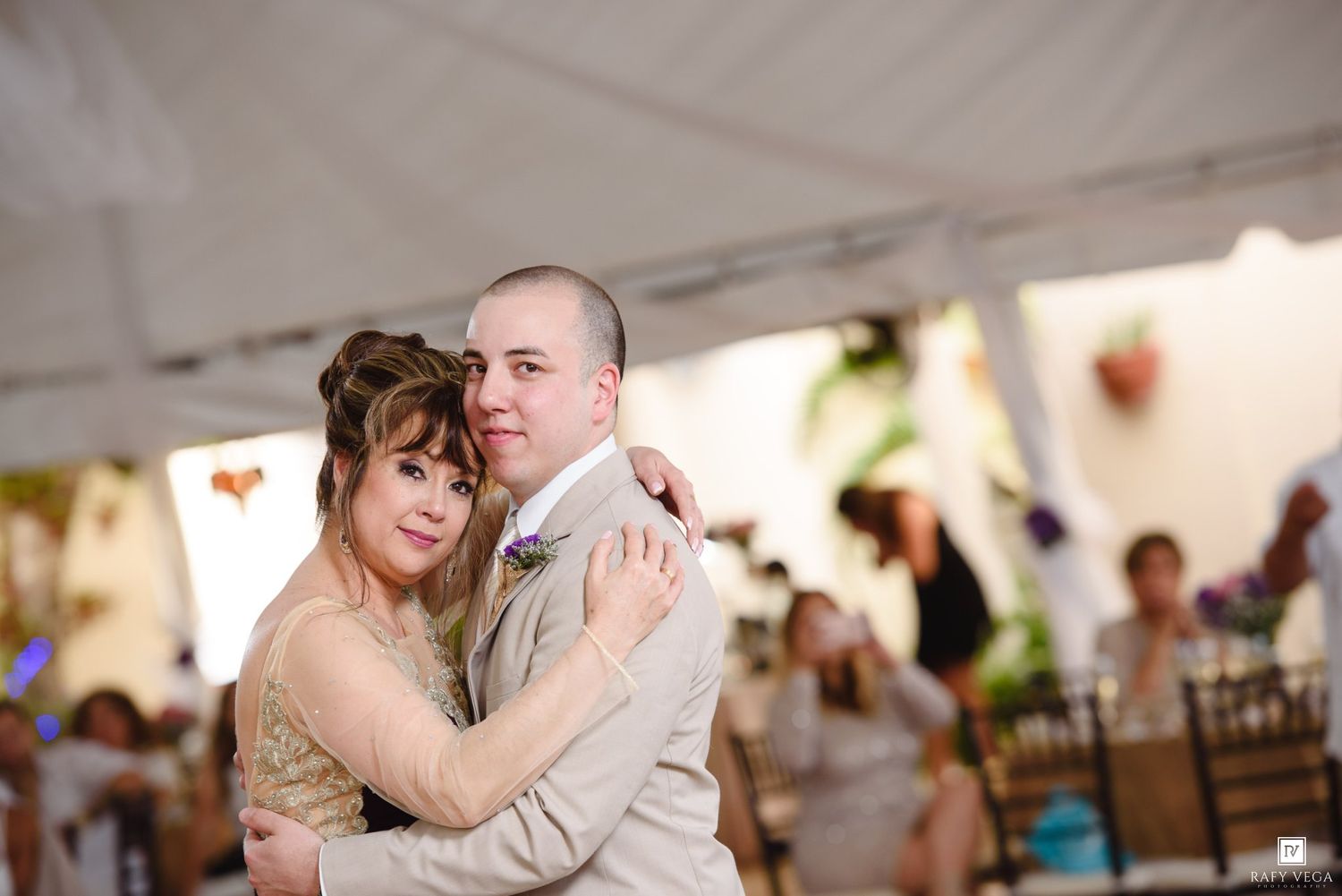 Couple shares first dance at wedding reception under white tent, groom in tan suit embracing bride in gold dress.