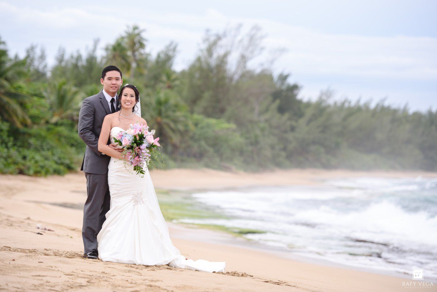 Bride and groom pose on a sandy beach with ocean waves and lush green trees in the background.