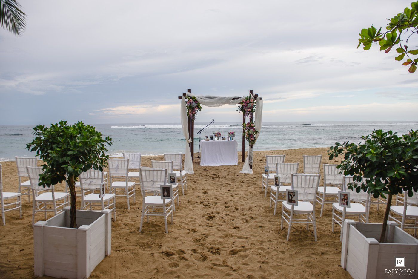 Beach wedding ceremony setup with white chiavari chairs, floral arch, and ocean backdrop on sandy shore.