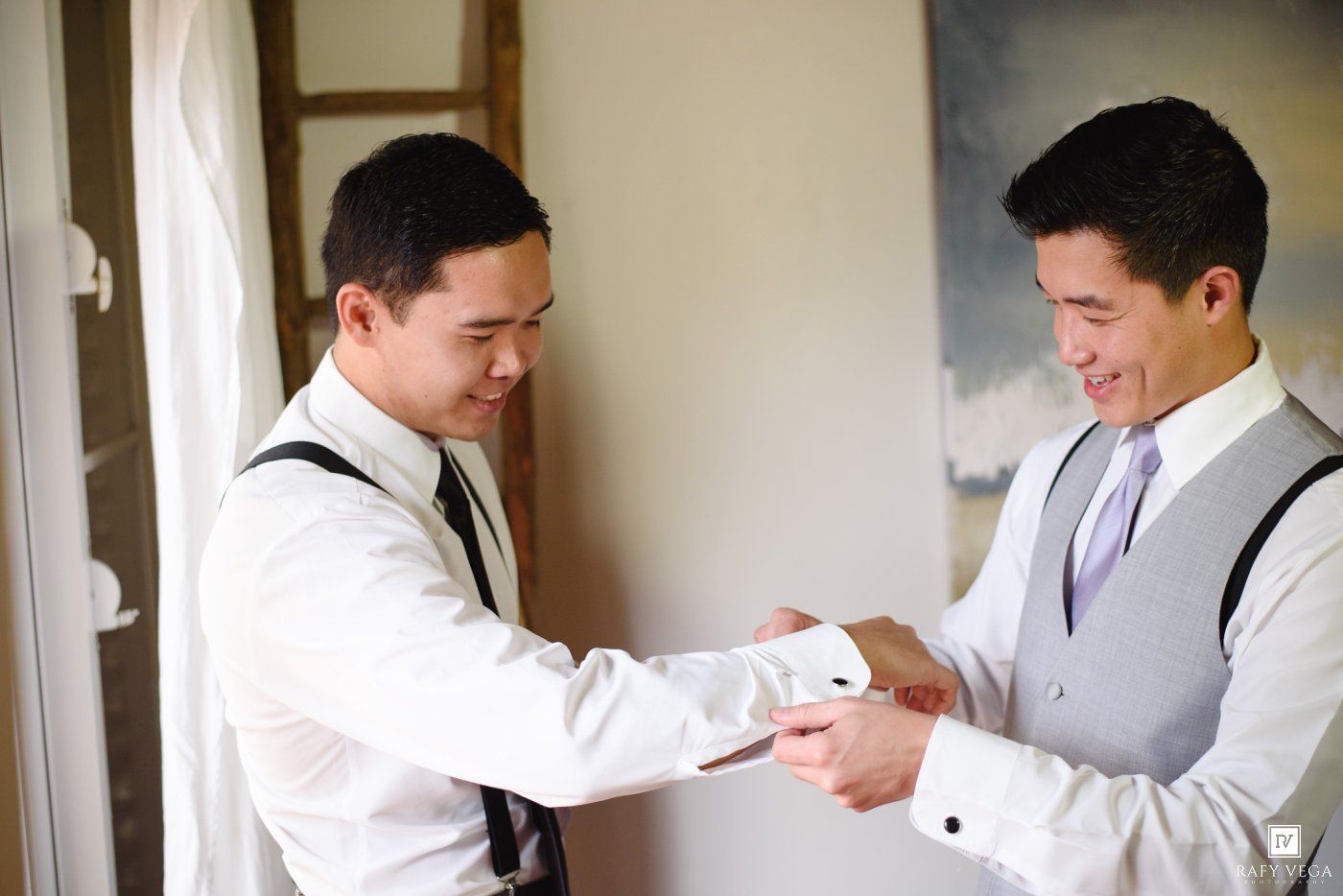 Two groomsmen in white shirts and suspenders adjusting cufflinks before a wedding ceremony.