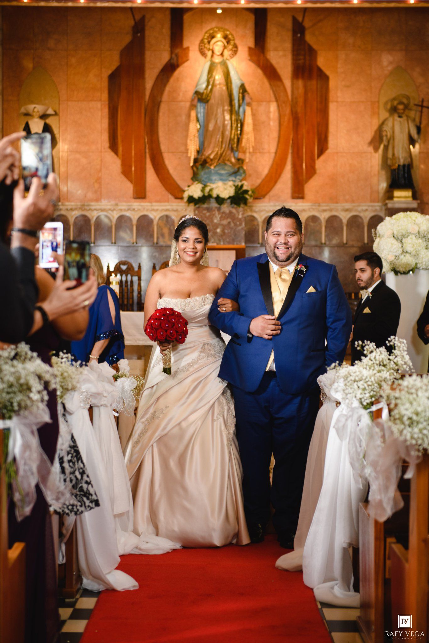 Bride and groom walk down church aisle after wedding ceremony, bride holding red bouquet, groom in blue suit.