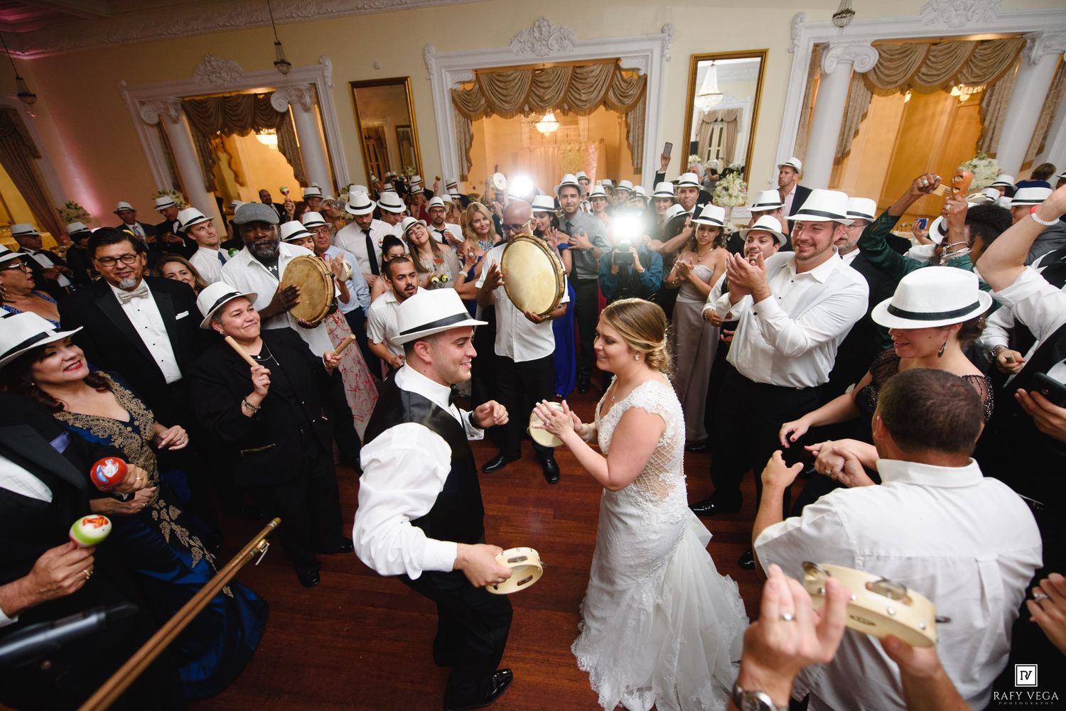 Bride and groom dance surrounded by guests in white fedora hats at elegant wedding reception ballroom celebration.