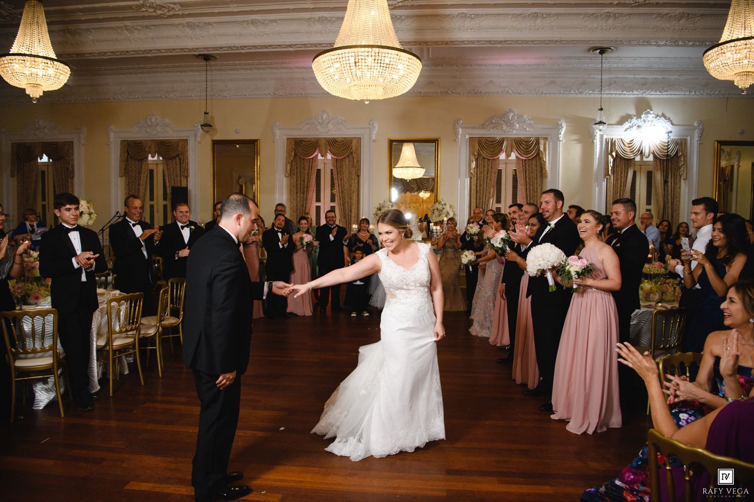 Bride and groom share first dance in elegant ballroom with chandeliers, surrounded by wedding guests and bridesmaids in pink.
