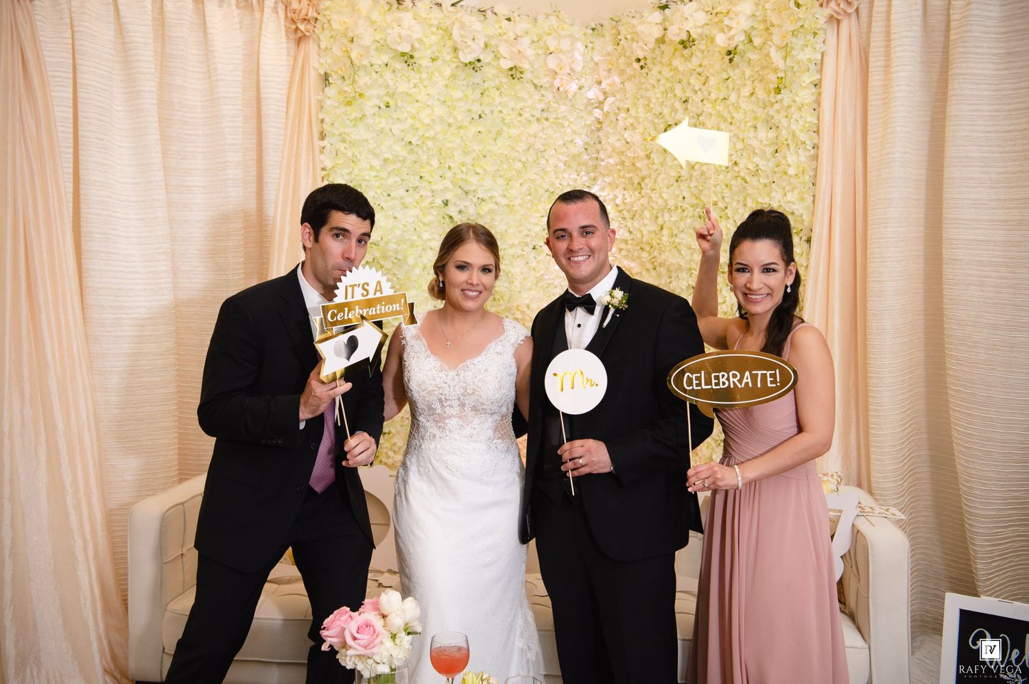 Four people posing at a wedding photo booth with fun props including a Celebrate sign and floral backdrop.
