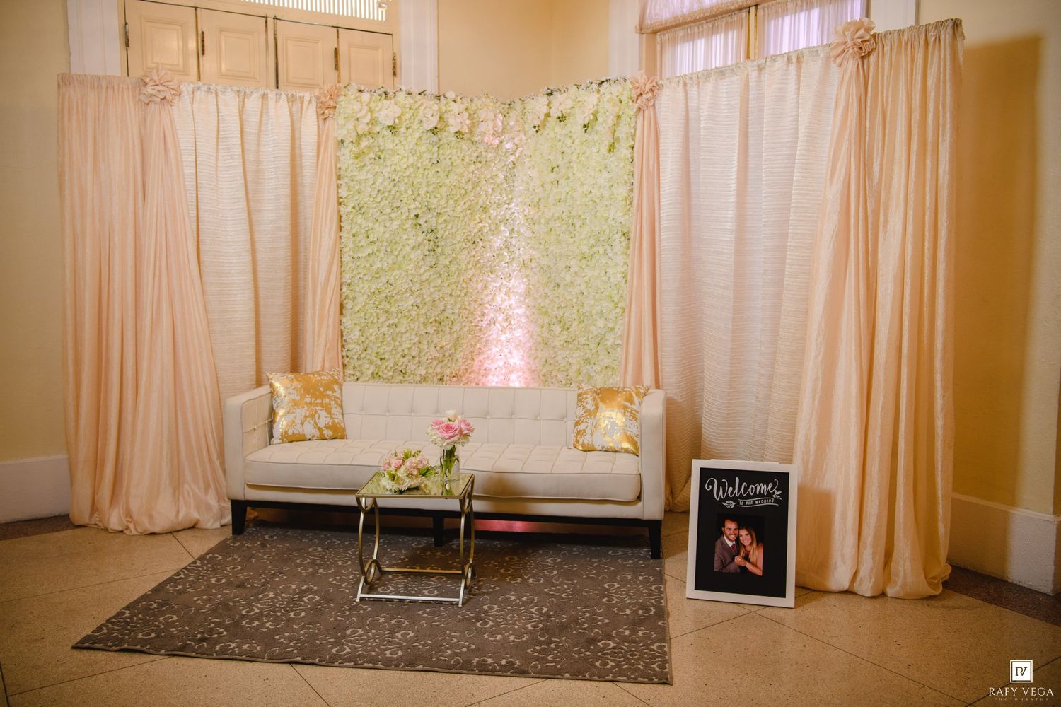 Elegant wedding reception lounge area with white sofa, floral wall backdrop, peach drapes, and welcome sign display.