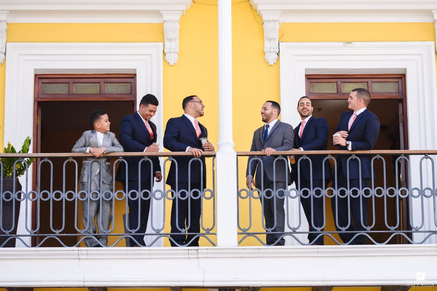 Groomsmen in navy suits with pink ties stand on a decorative balcony of a yellow building.