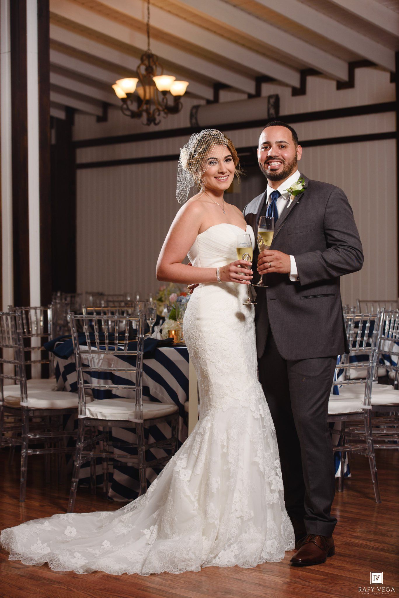 Bride in white lace mermaid gown and groom in gray suit toasting champagne at elegant wedding reception.