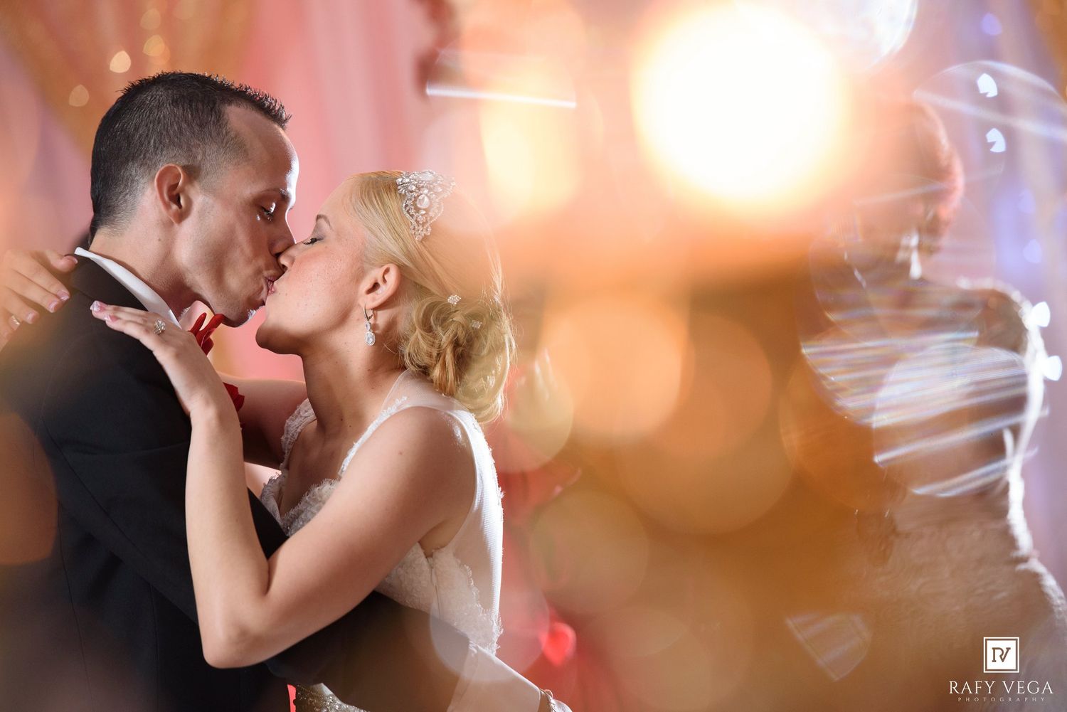 Bride and groom share a romantic first kiss on the dance floor surrounded by warm bokeh lights at their wedding reception.