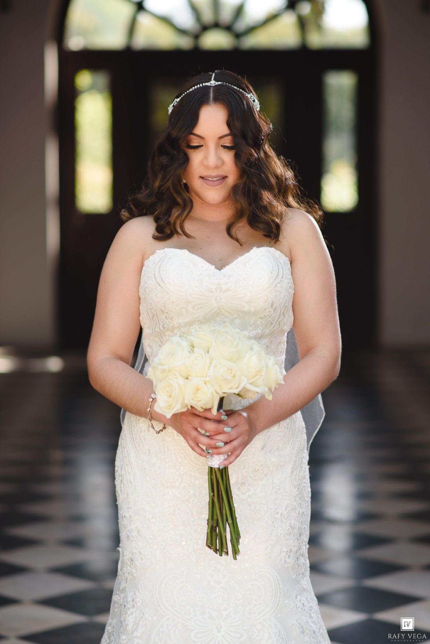Bride in strapless lace gown holding white rose bouquet, wearing veil and headpiece, standing in elegant venue.