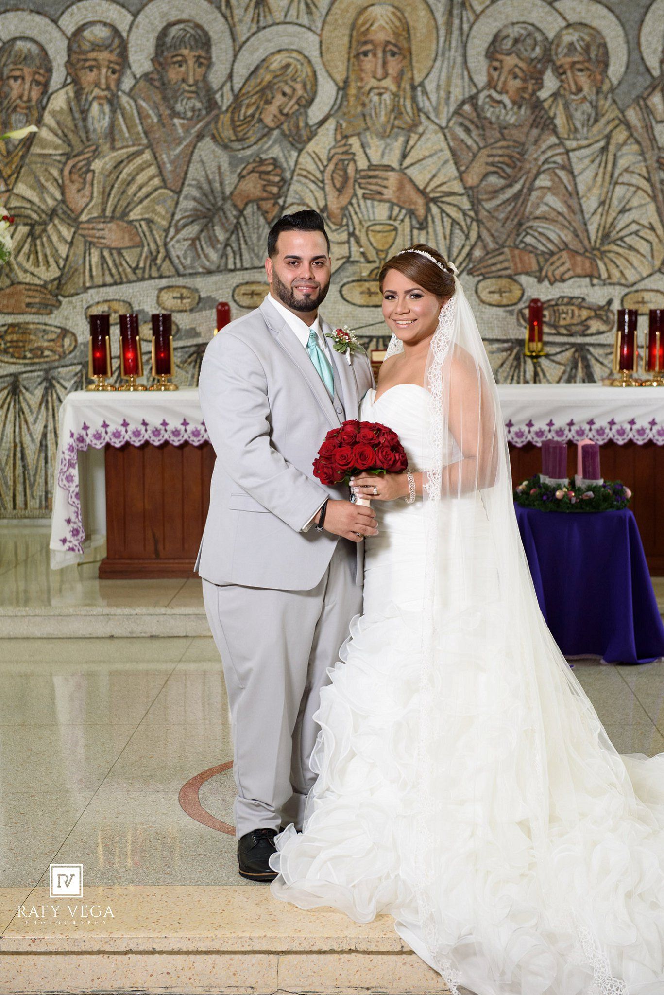 Bride and groom posing at church altar, bride holding red rose bouquet in white ruffled gown with veil.