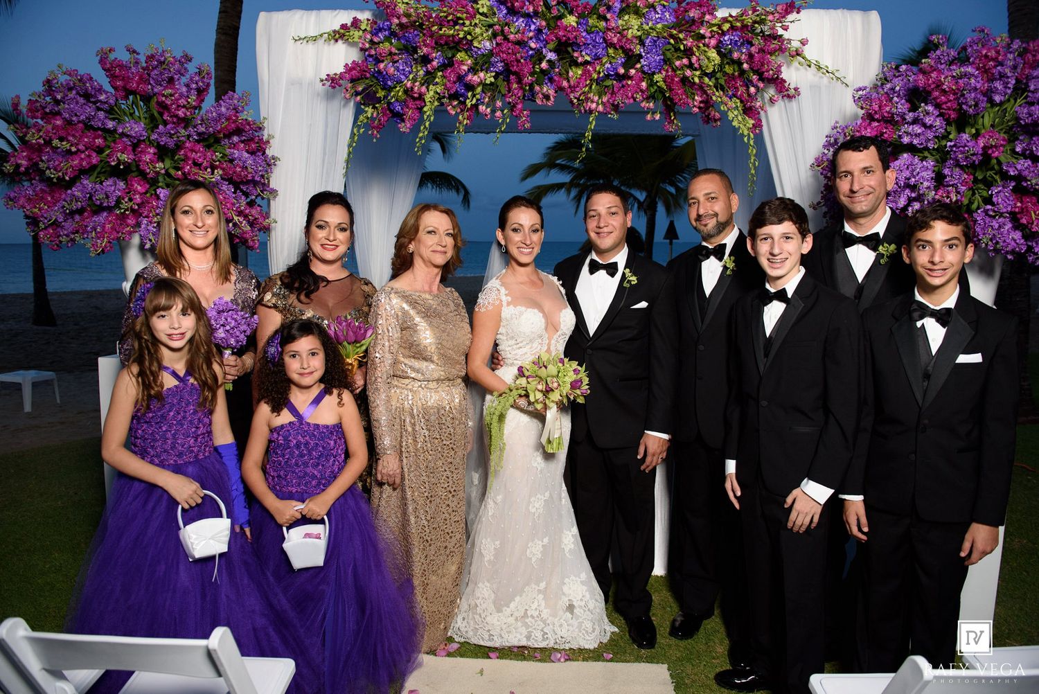Wedding party poses under floral arch with purple flowers, bride in white gown, groom in tuxedo, family in formal attire.