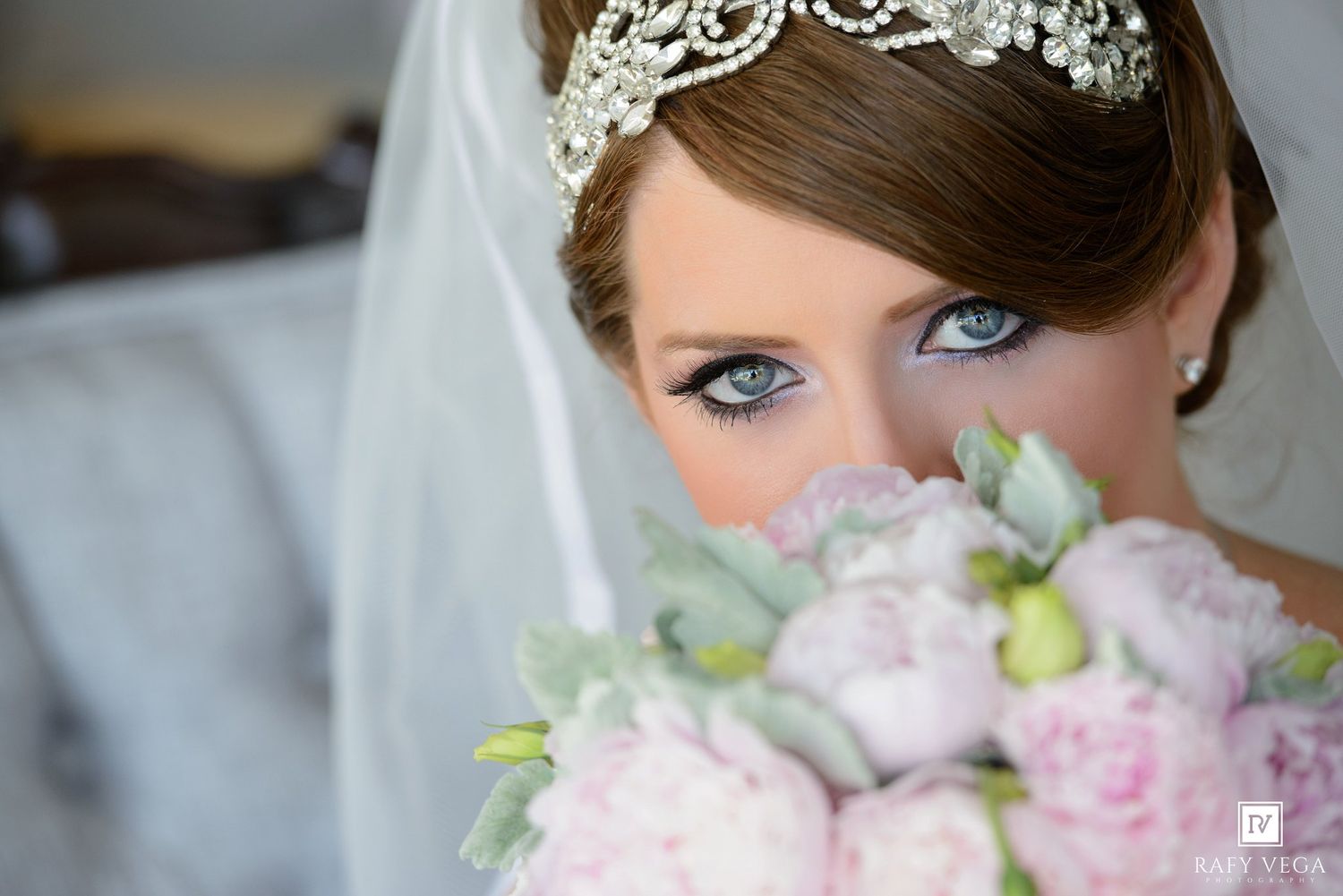 Bride with blue eyes wearing a crystal tiara and veil holds pink peony bouquet close to her face.
