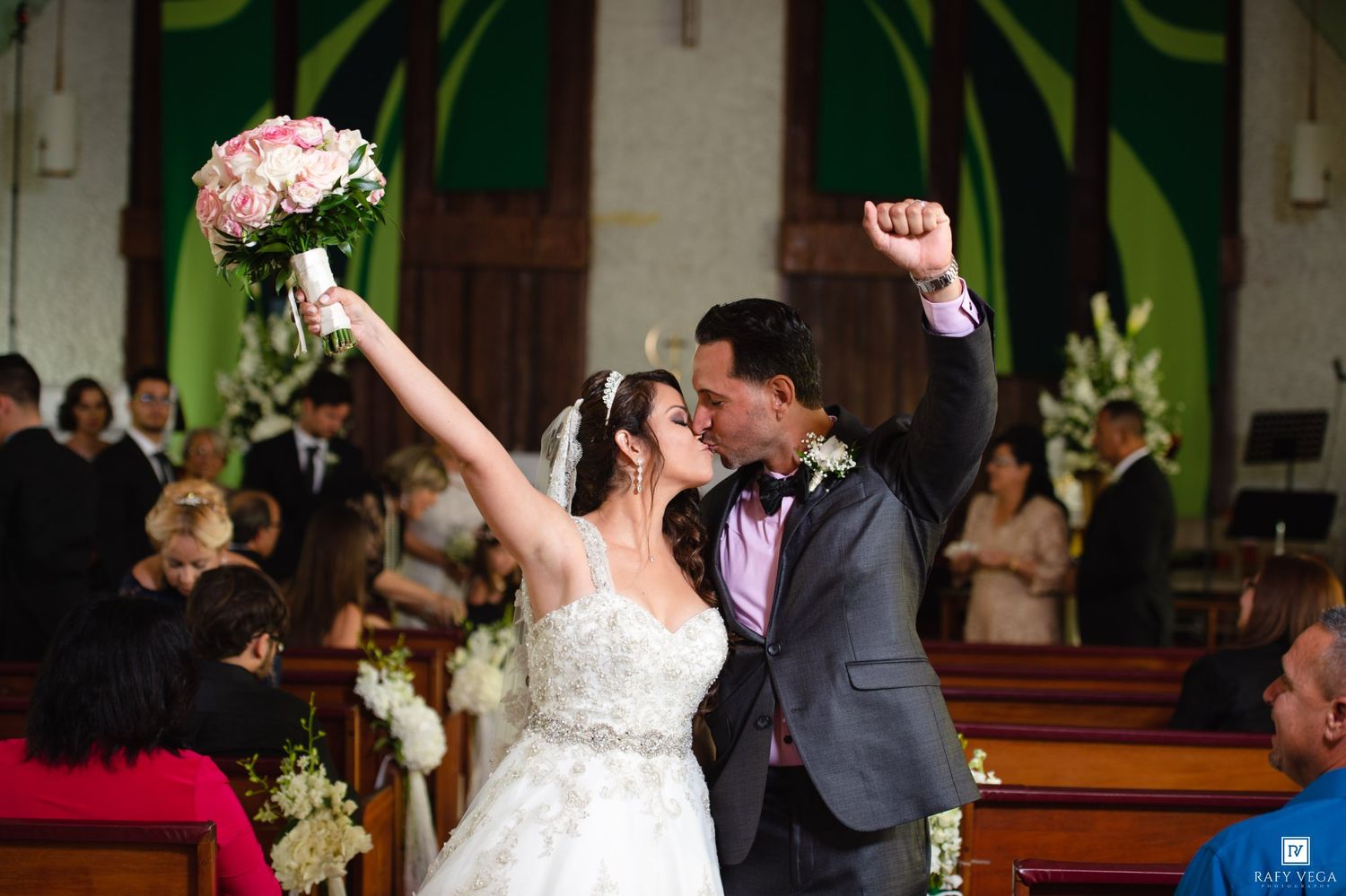 Joyful newlyweds celebrate at church altar, bride in white gown holding pink bouquet aloft, groom in gray suit, arms raised.