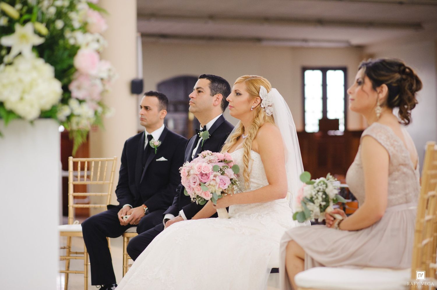 Bride and groom sit at wedding ceremony altar with bouquet of pink roses, flanked by groomsman and bridesmaid in elegant attire.
