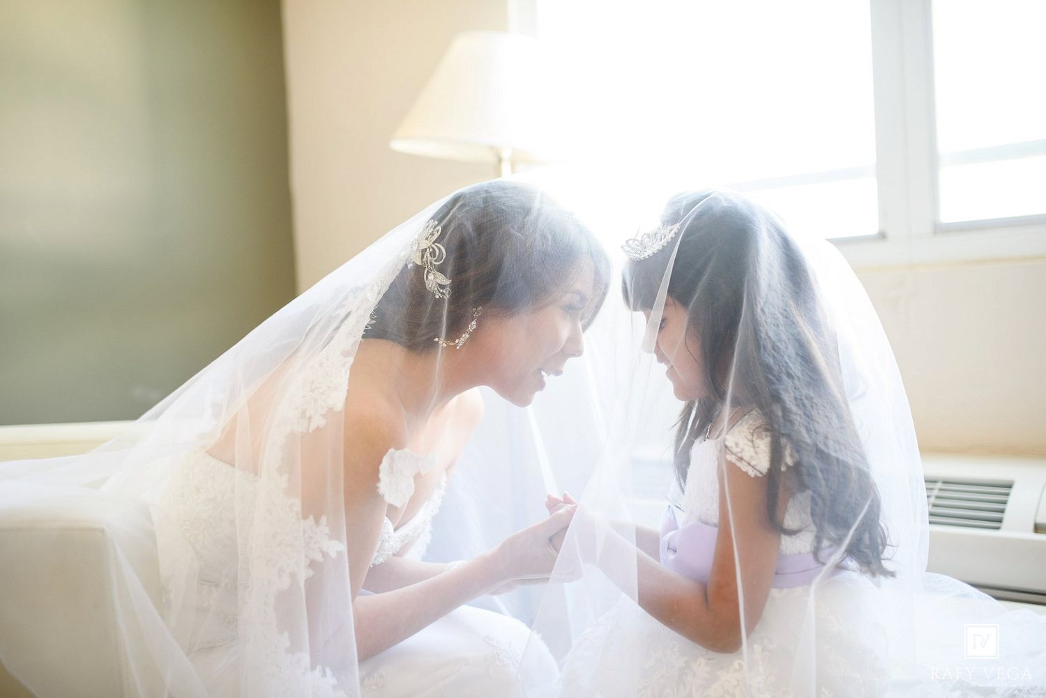 Bride and flower girl share an intimate moment under white veils, sitting together in soft natural light.