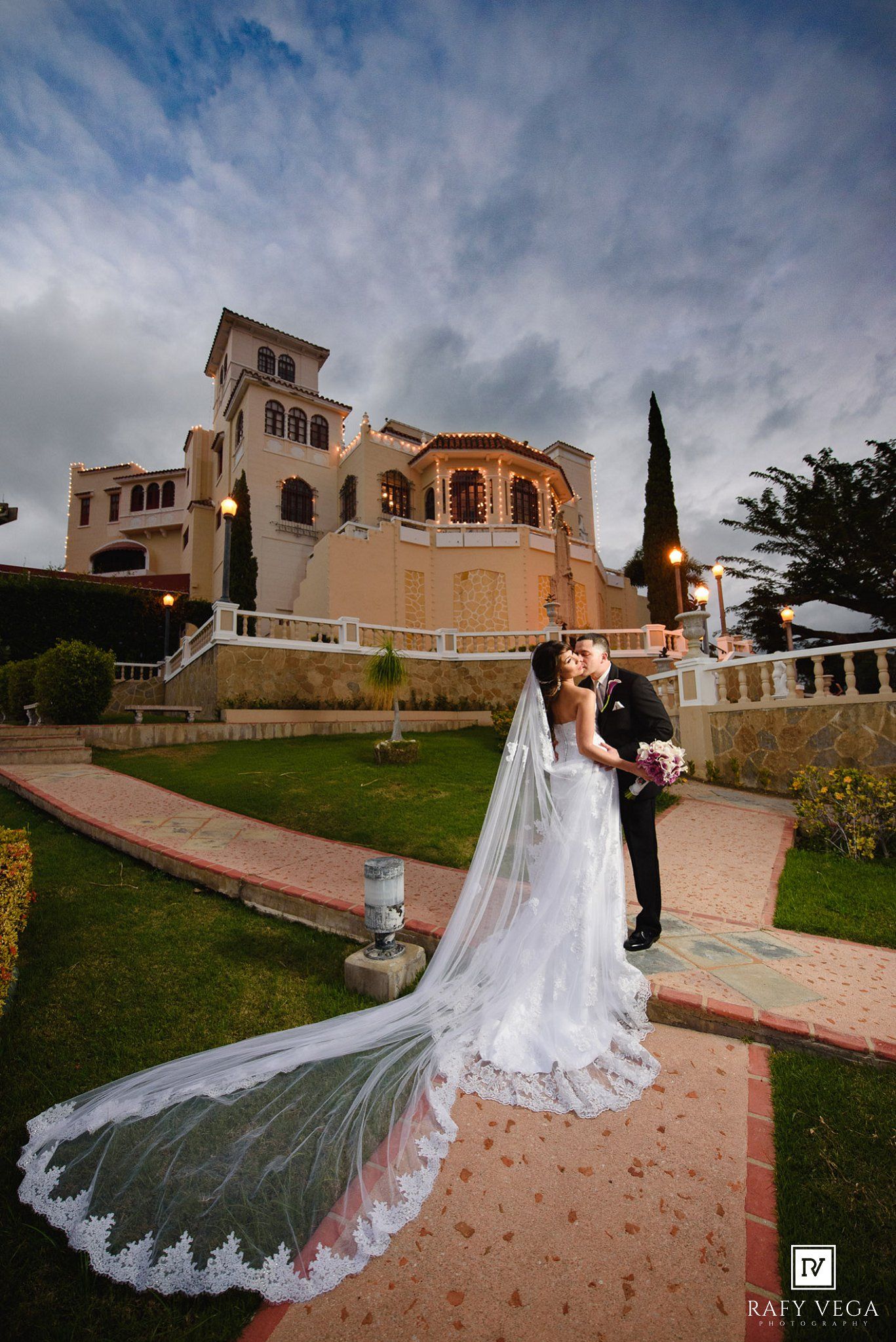 Bride and groom share a romantic moment outside an elegant villa at dusk, long lace veil trailing.