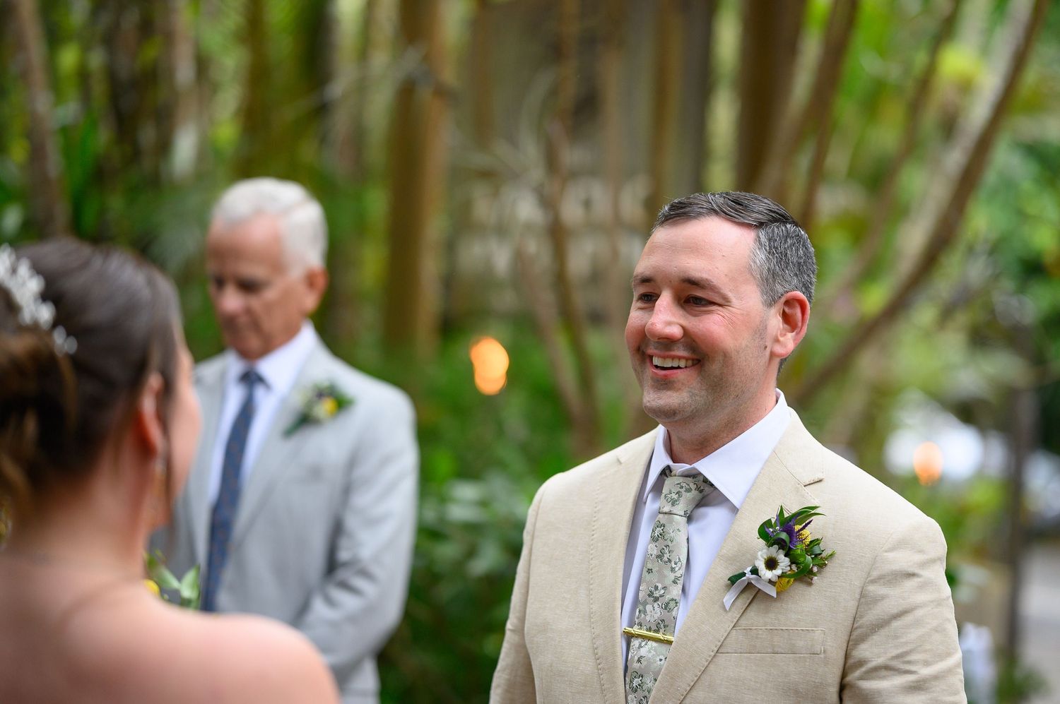 Smiling groom in tan suit with floral boutonniere during outdoor wedding ceremony surrounded by lush greenery.