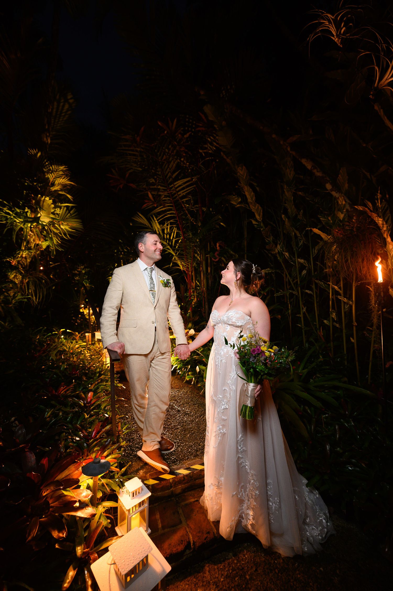 Bride and groom holding hands at night in a tropical garden, illuminated by warm lantern light.