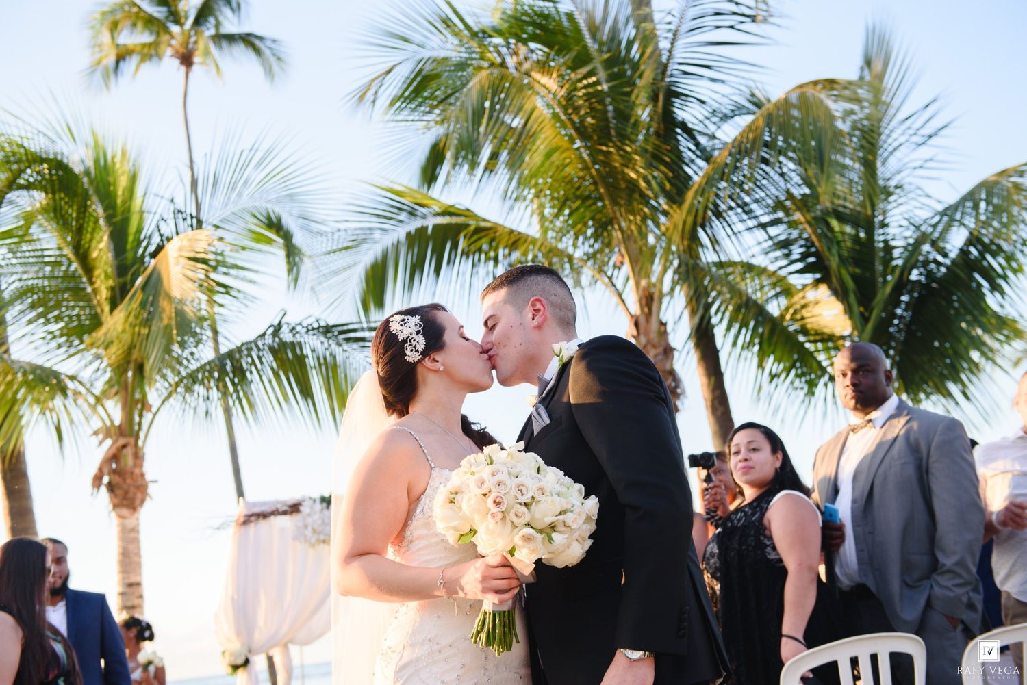 Bride and groom share a kiss at outdoor beach wedding ceremony with palm trees in tropical destination.