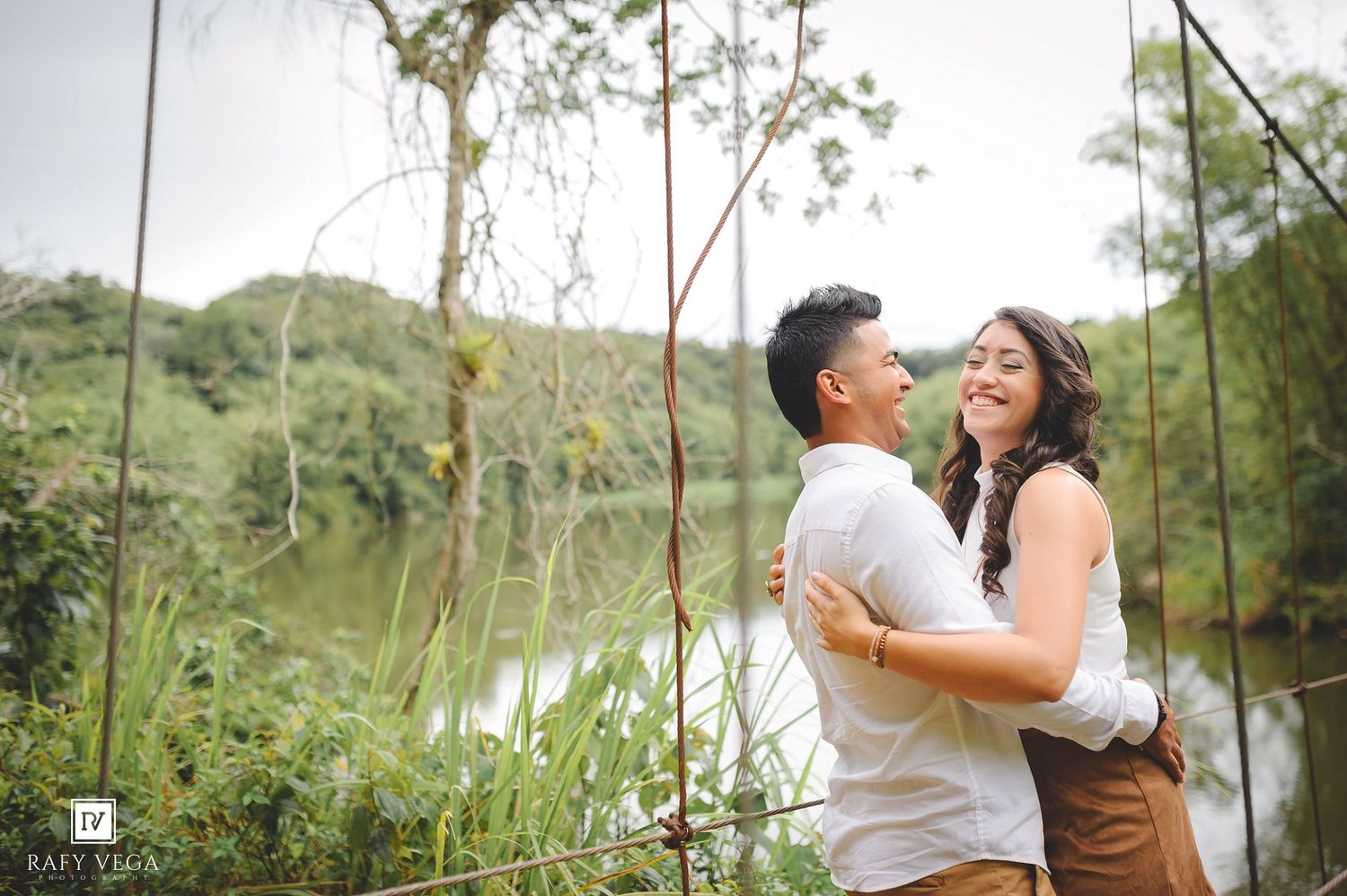Couple embracing and smiling near a serene lake surrounded by lush greenery during an outdoor engagement photo session.