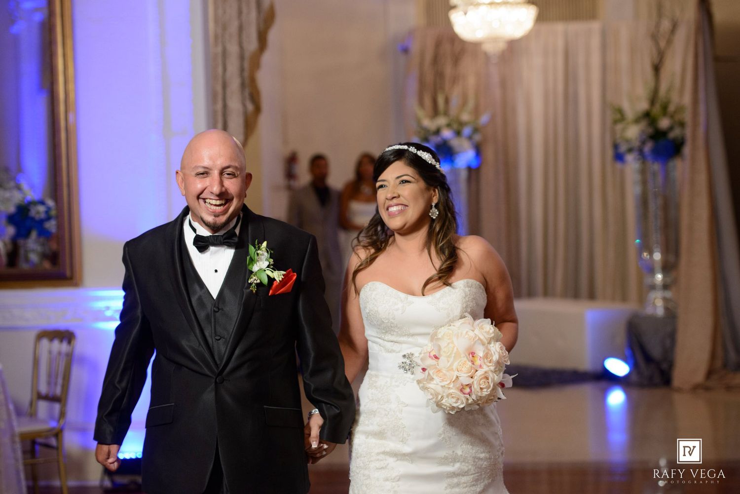 Smiling bride in white strapless gown holding bouquet walks with groom in black tuxedo at elegant wedding reception.