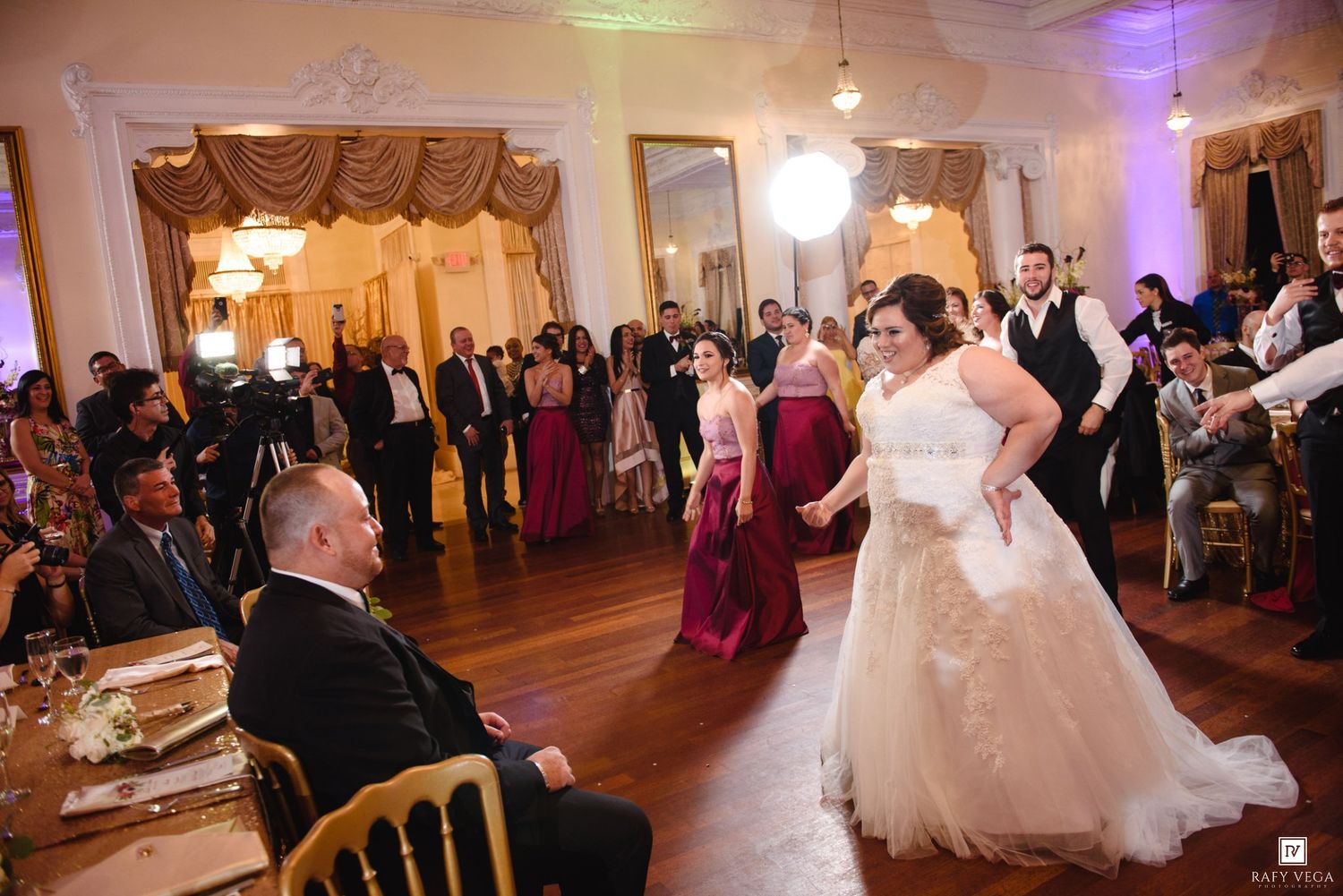 Bride in white gown performs first dance while groom sits watching in elegant ballroom with guests gathered around.