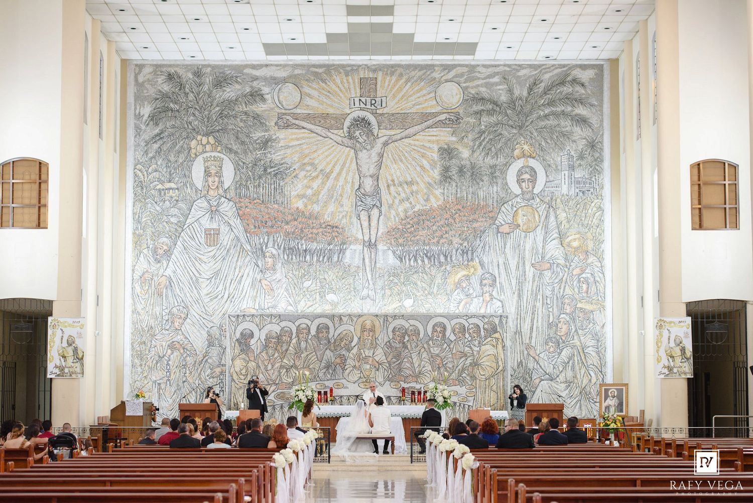 Catholic church interior with large mosaic of crucifixion, wedding ceremony with guests seated in wooden pews.