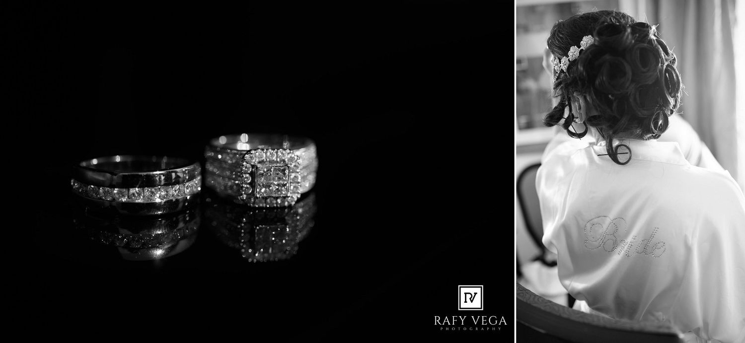 Black and white wedding rings and diamond engagement ring displayed on reflective surface beside bride preparing in white gown.