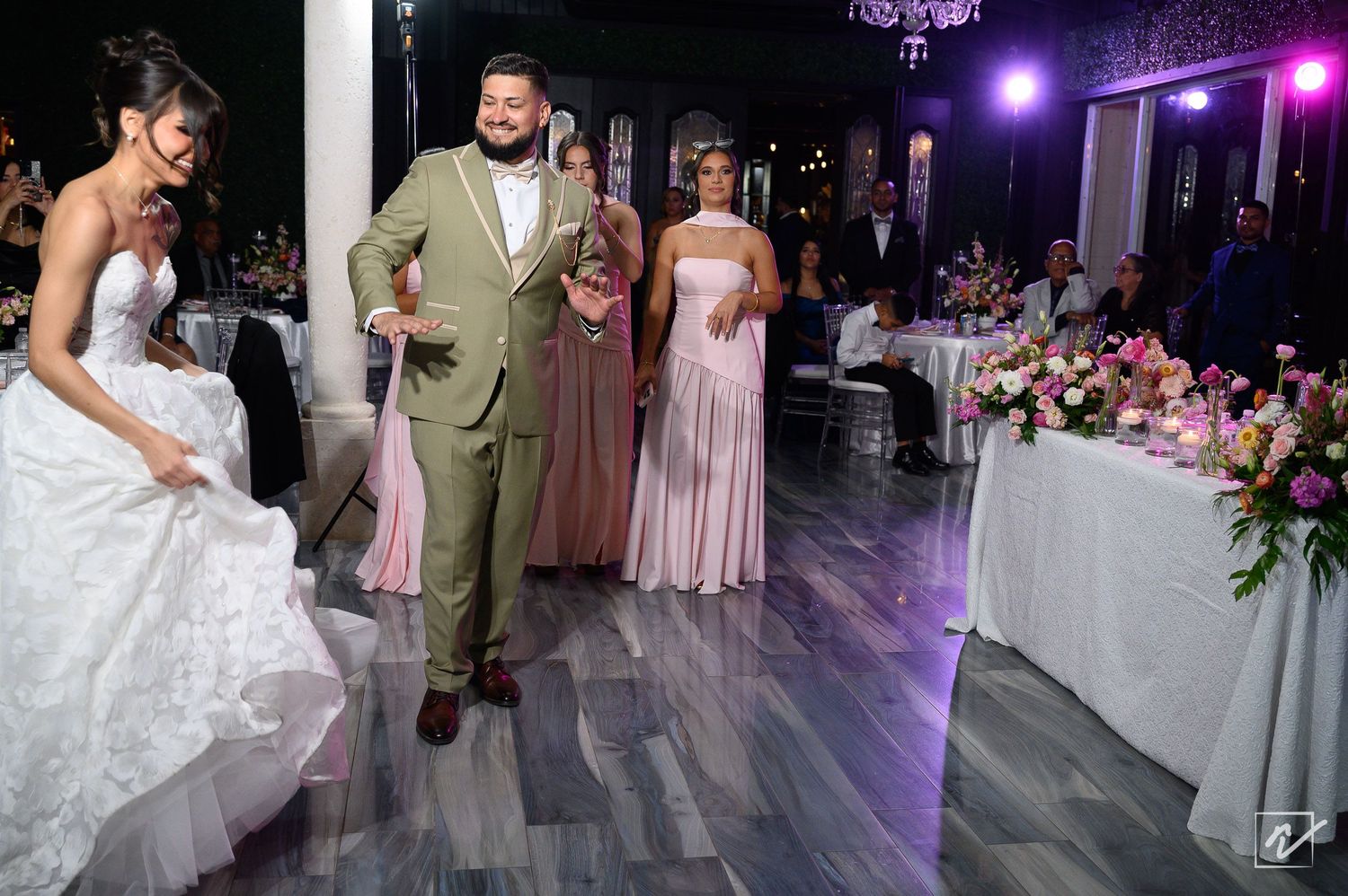 Bride in white gown and groom in olive suit walk across a reception dance floor as bridesmaids in blush dresses stand near floral tables.