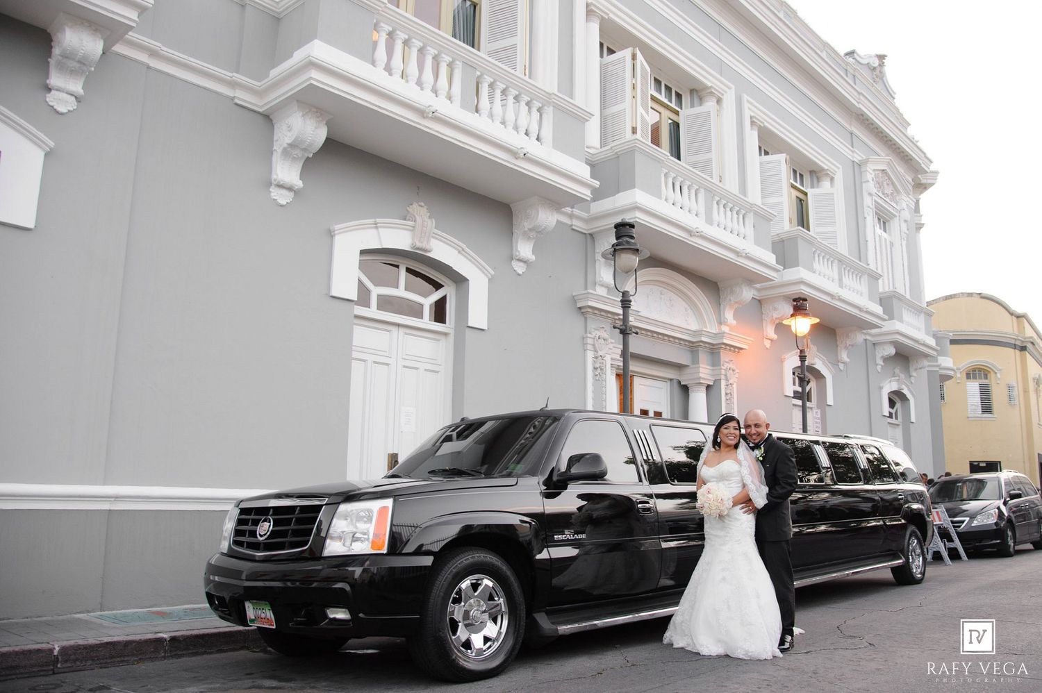 Bride and groom posing in front of a black stretch Cadillac Escalade limousine outside an elegant white building.