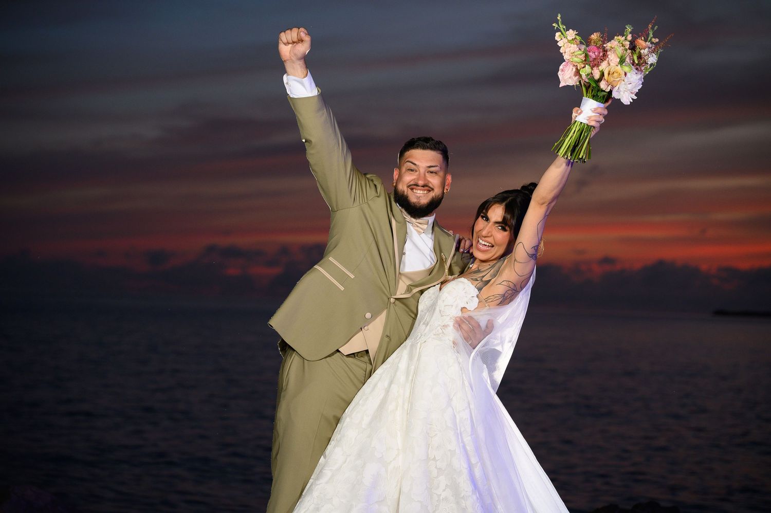 Joyful newlyweds celebrate at sunset by the ocean, bride in white gown holding bouquet, groom in tan suit raising fist.