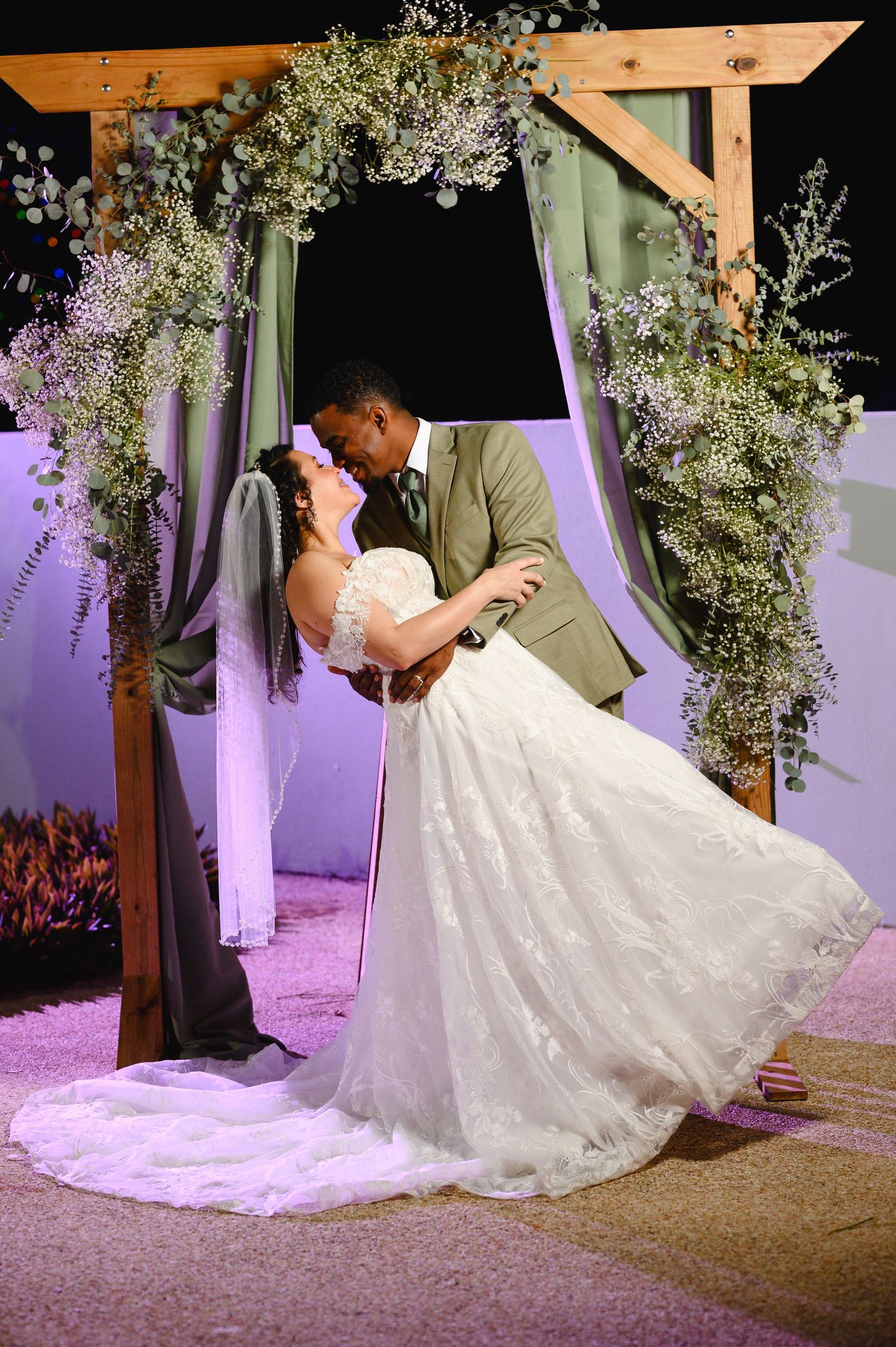 Bride and groom share a romantic dip kiss under a wooden arch adorned with greenery and flowers at their wedding.