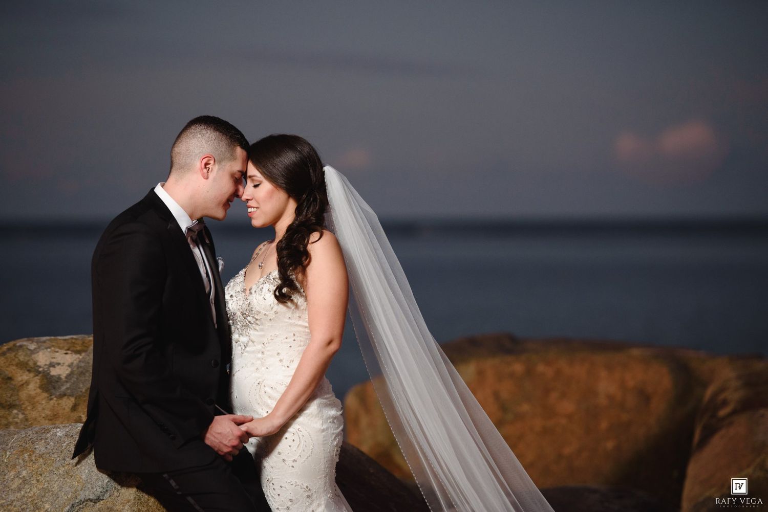 Bride and groom share romantic moment on rocky shoreline at sunset, bride wearing lace gown and veil.