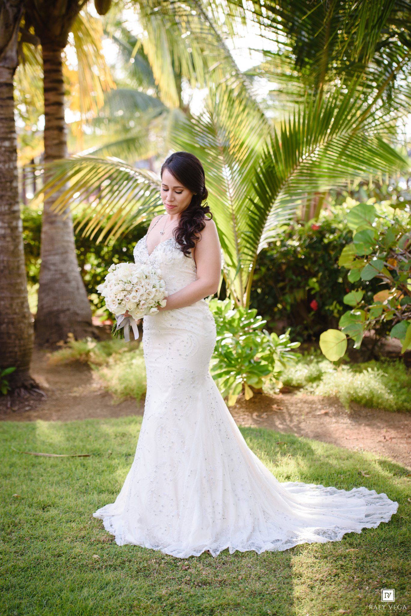 Bride in white lace mermaid gown holding bouquet of white flowers, standing in tropical garden with palm trees.