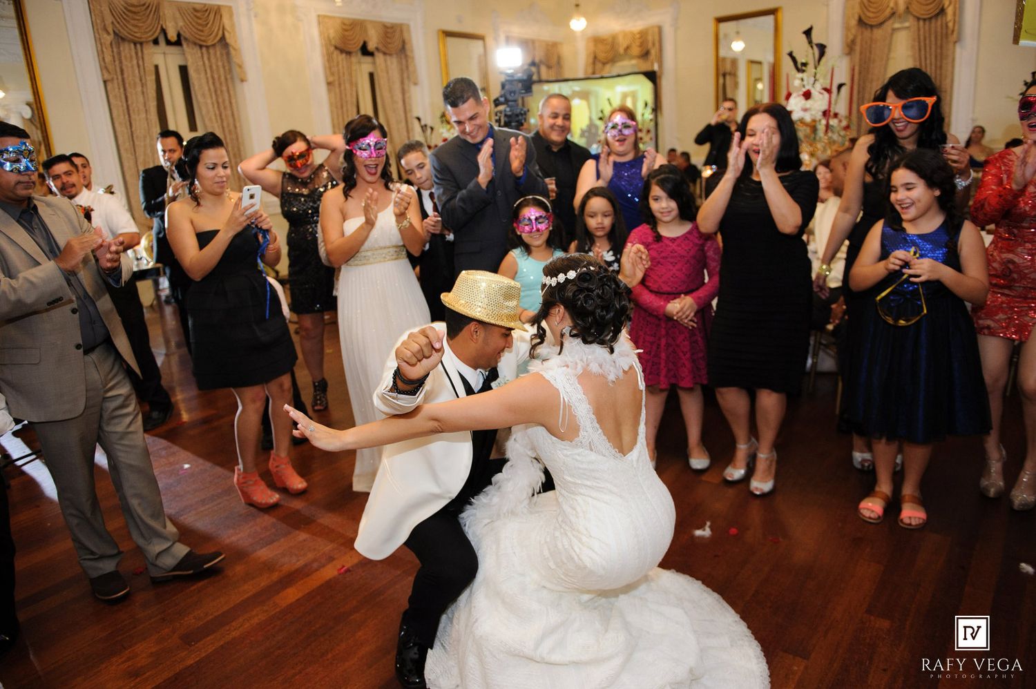Bride and groom dancing joyfully at wedding reception while guests cheer and photograph the celebration.