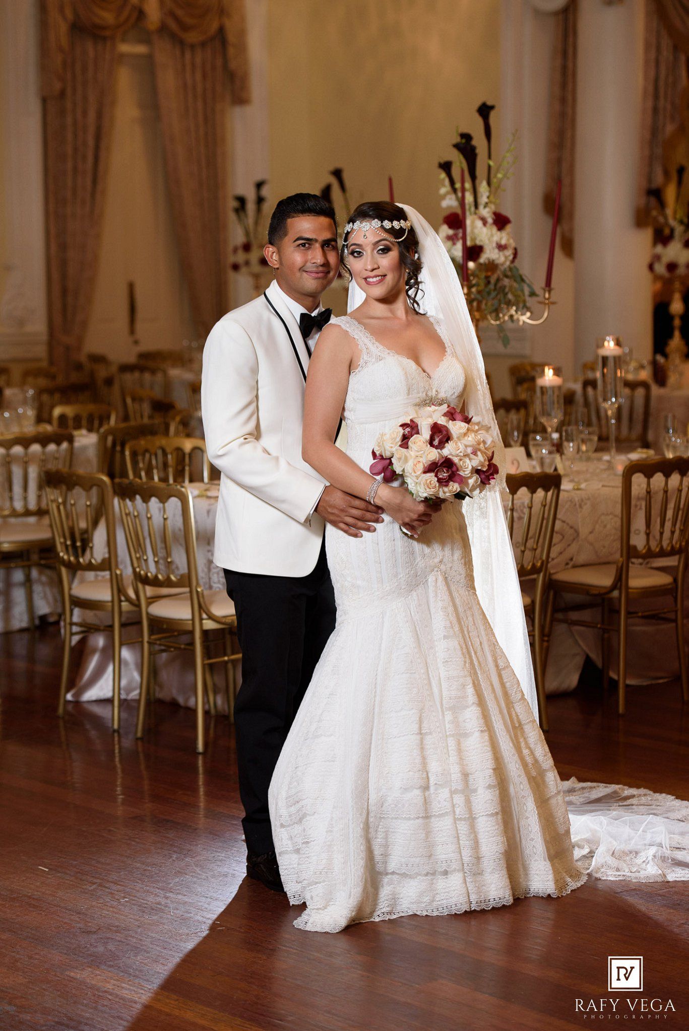Elegant bride and groom posing in a grand ballroom, bride in mermaid gown holding bouquet, groom in white tuxedo jacket.