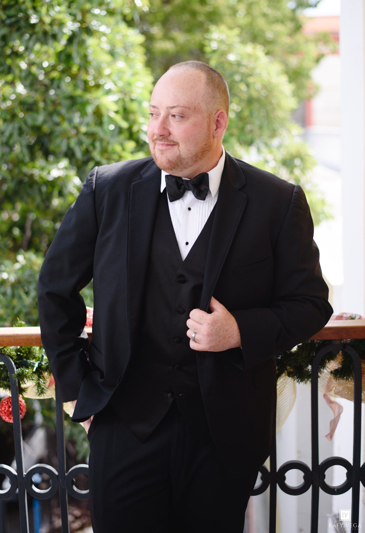A well-dressed man in a black tuxedo with bow tie poses on a decorated balcony surrounded by lush greenery.