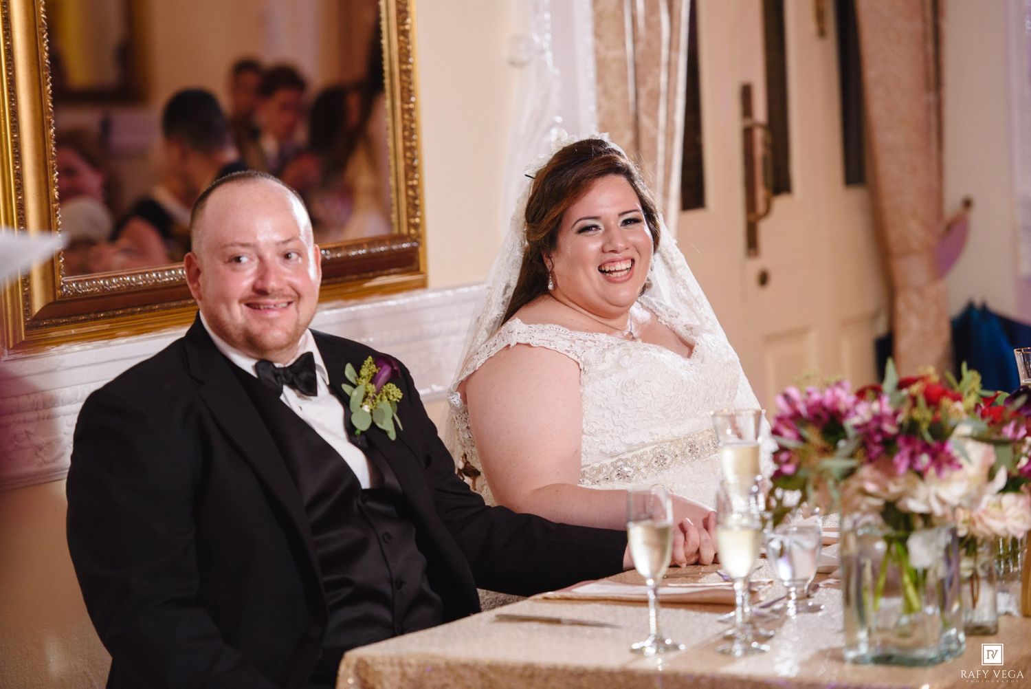 Bride and groom smiling at wedding reception sweetheart table with champagne and floral centerpiece.