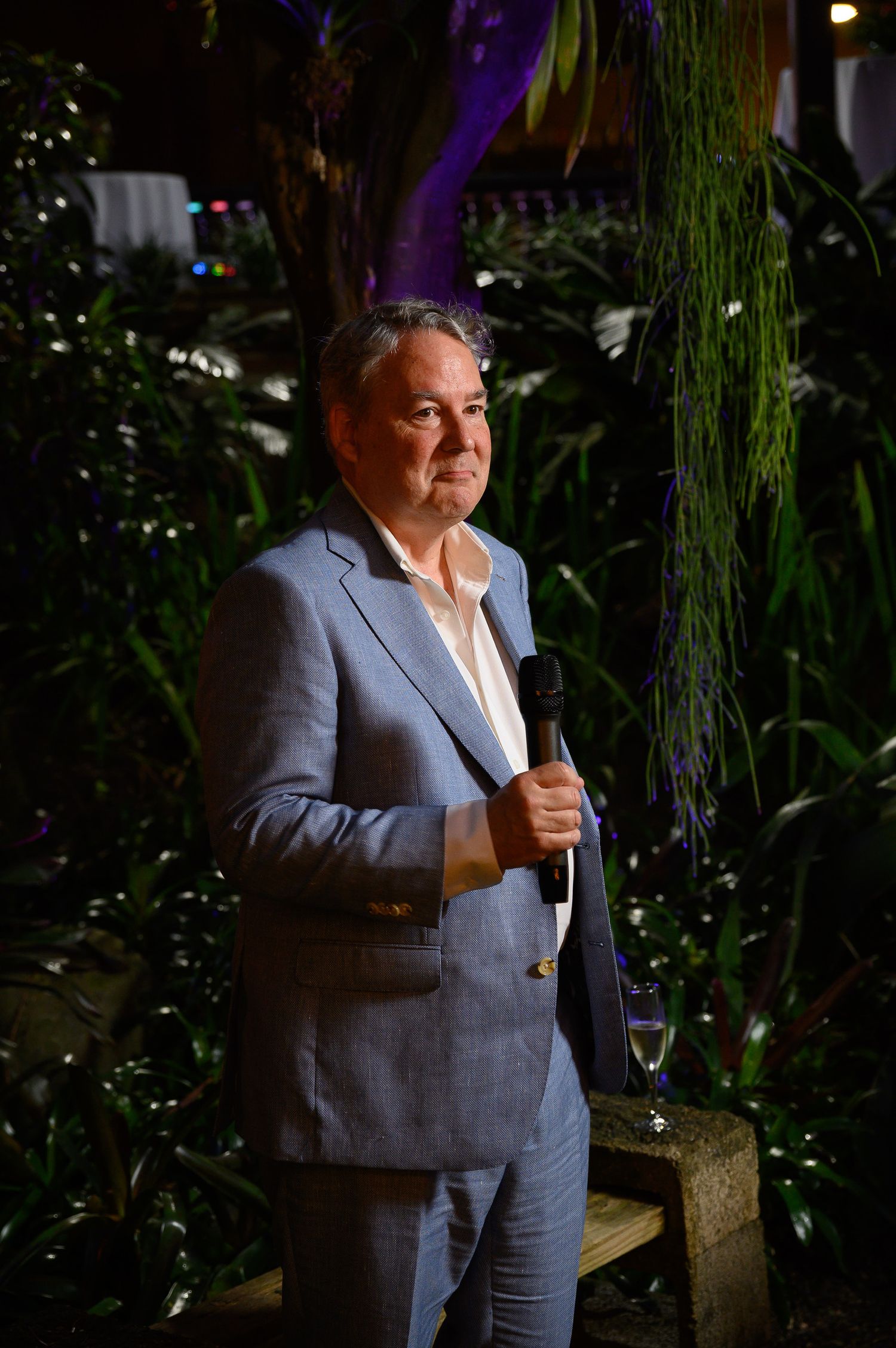 Man in light blue suit holding microphone speaks at elegant indoor garden event at night.