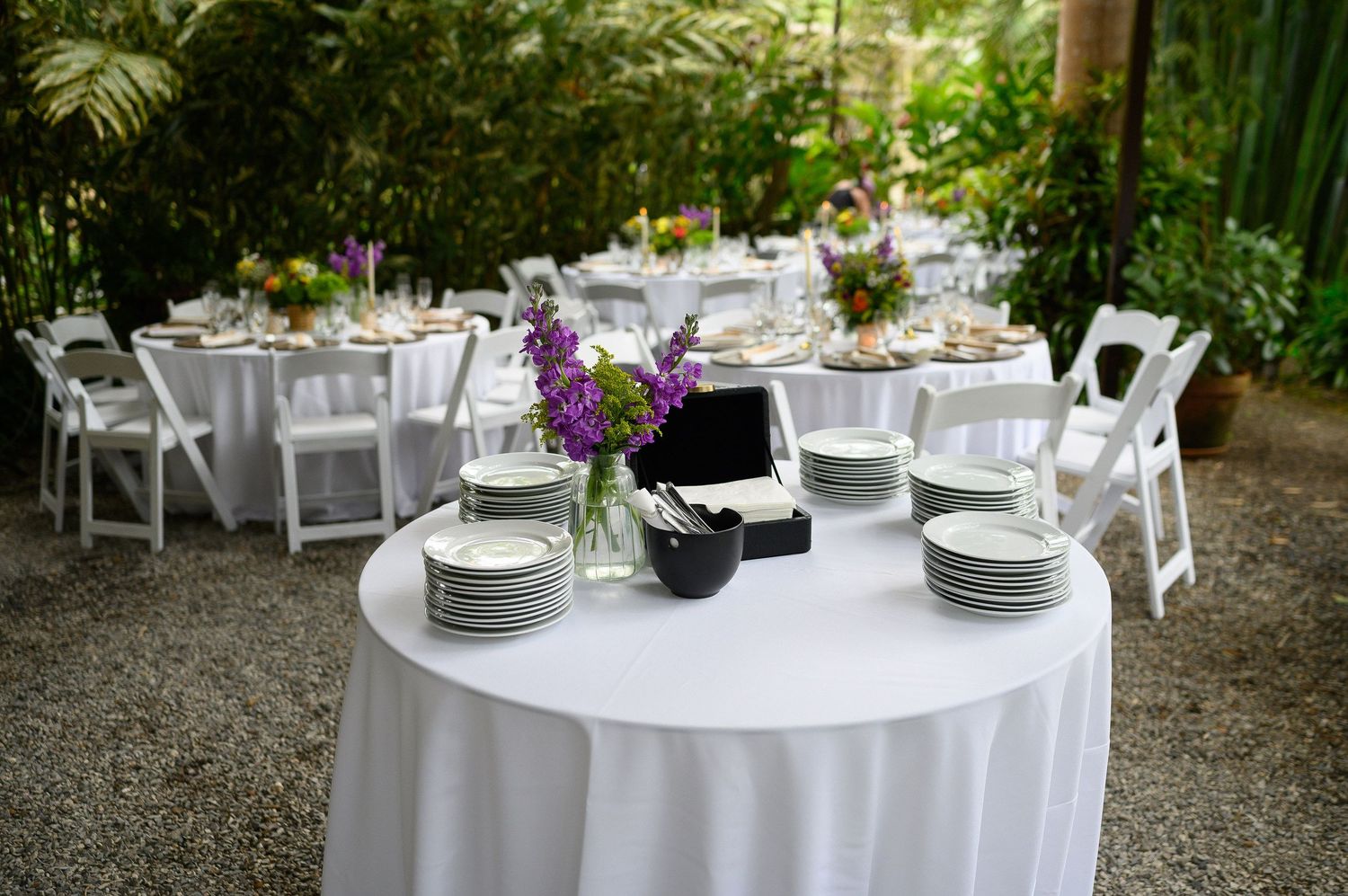 Elegant outdoor wedding reception with white round tables, folding chairs, purple floral centerpieces, and lush green tropical backdrop.