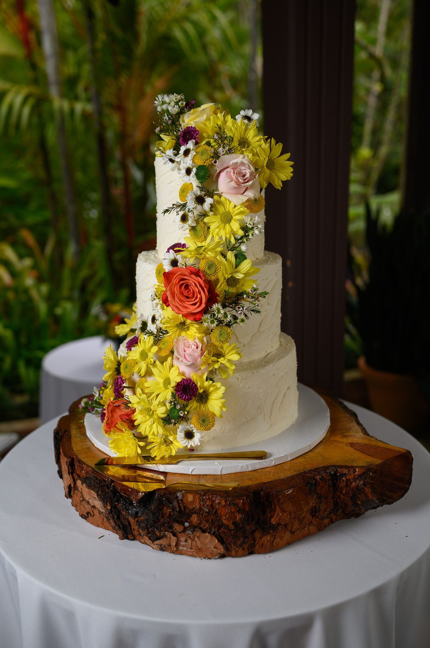 Four-tier white wedding cake adorned with yellow daisies, pink and orange roses on a rustic wood slice stand.
