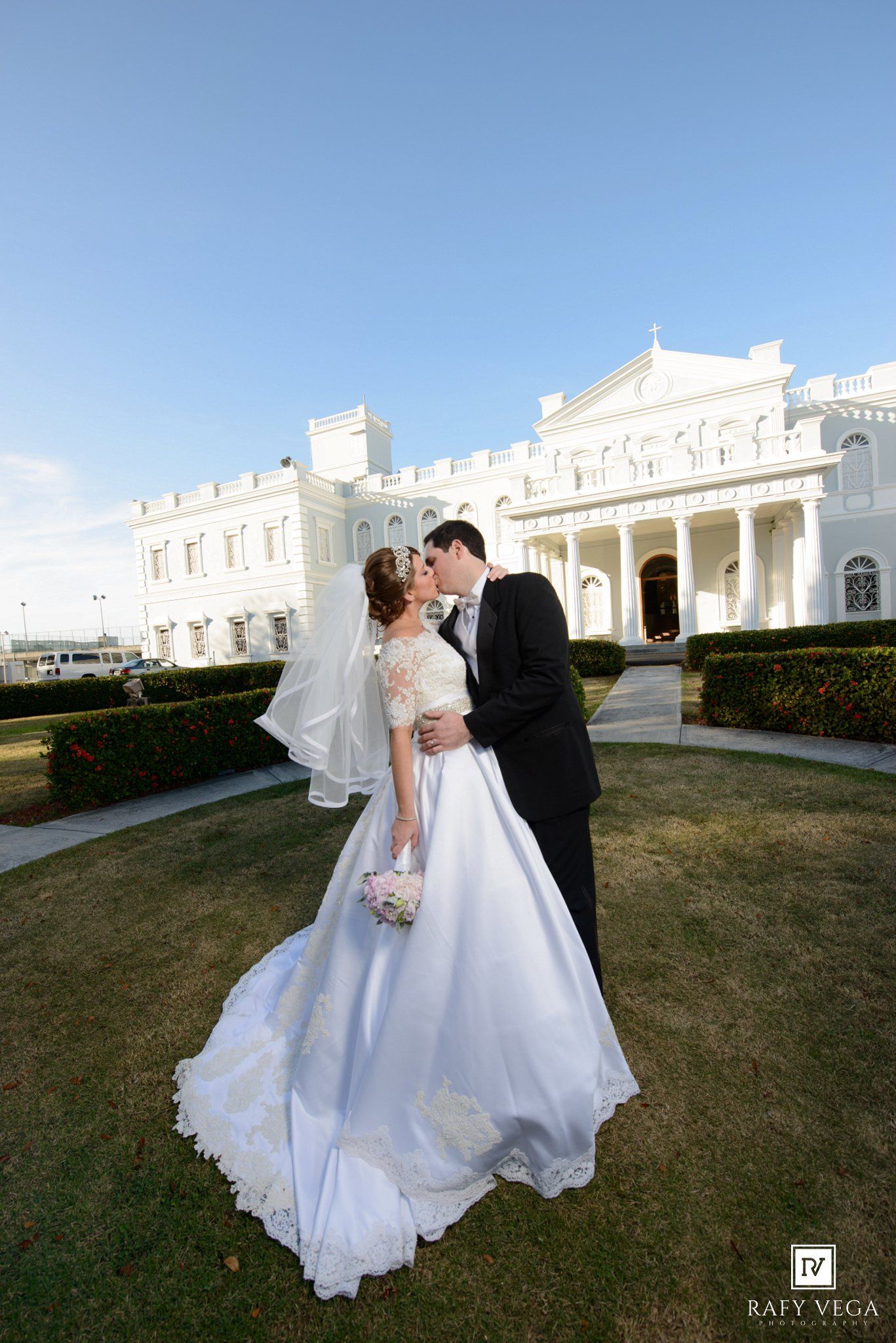 Newlyweds share a kiss in front of a grand white mansion, bride in a flowing lace wedding gown.